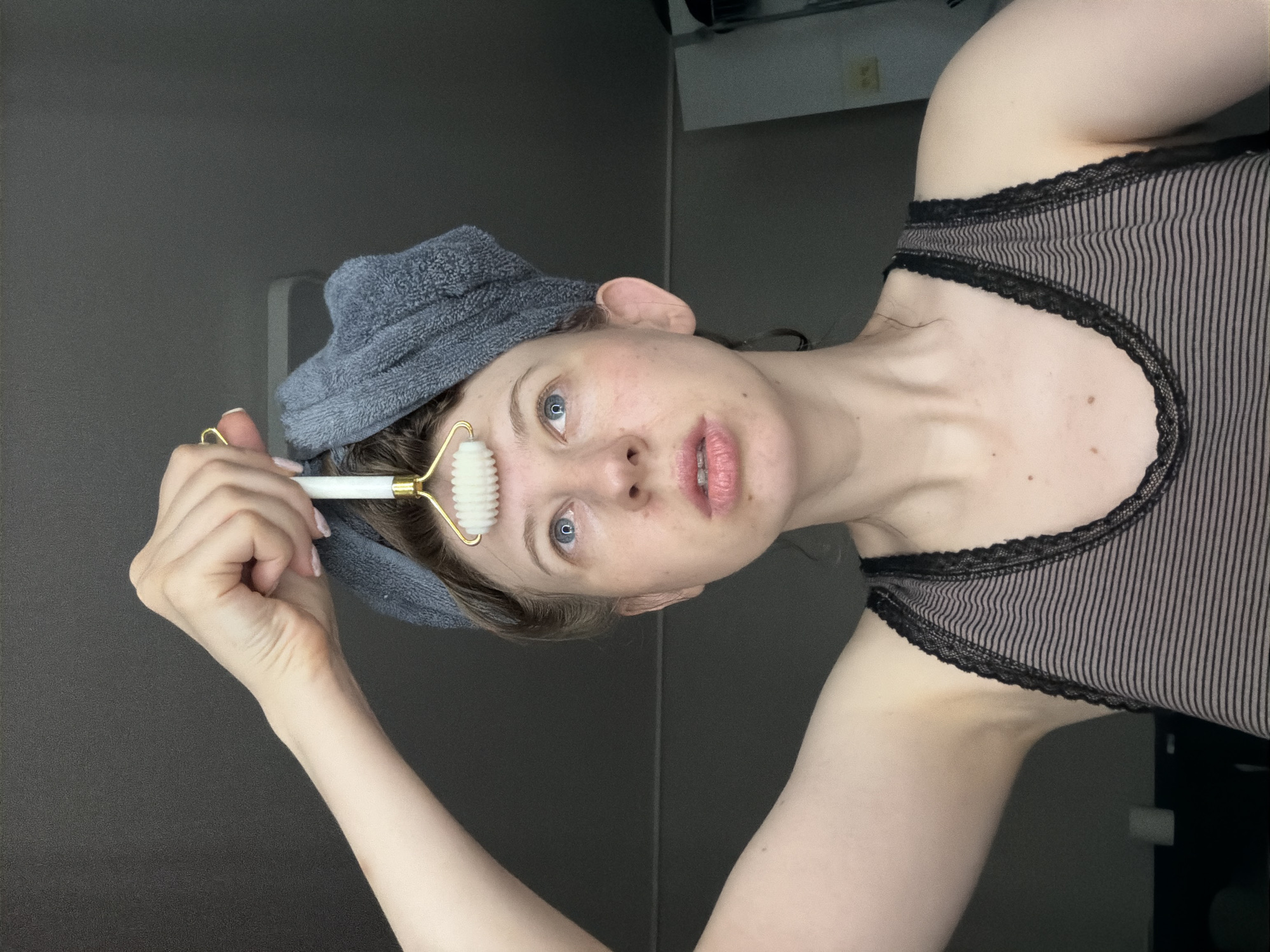 A woman with a gray towel wrapped around her head is holding a white jade roller to her forehead. She is wearing a striped tank top. The background is a dark, plain wall.