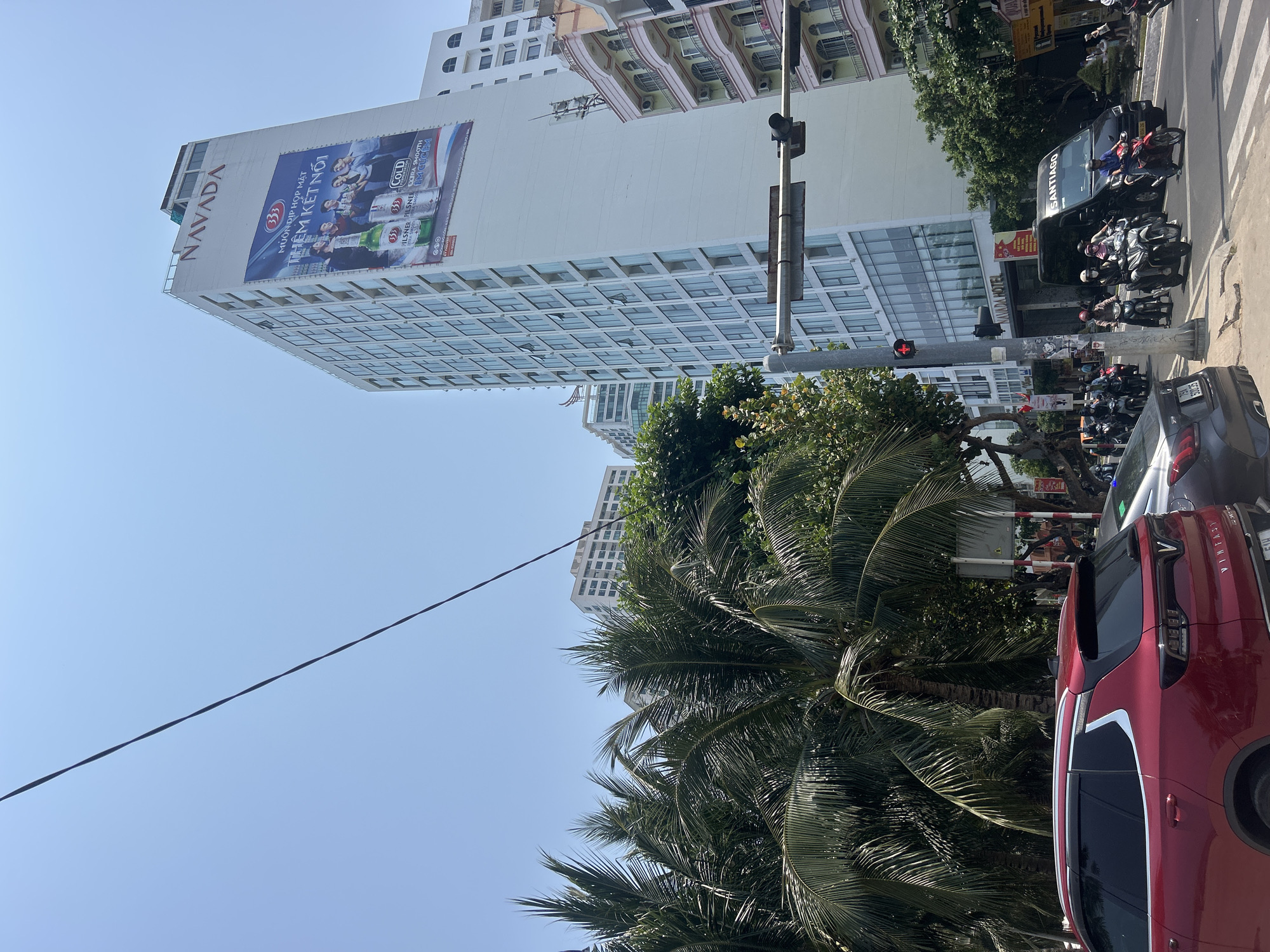 The image shows a street scene in Bắc Nha Trang, Vietnam. A tall, modern building with a large billboard advertising "333" beer dominates the left side of the frame. The building is identified as "NAVADA HOTEL."
The street is busy with traffic, including motorbikes, cars, and a van. A red SUV is prominently positioned on the right side of the image. A traffic light is visible, showing a red light. Tropical vegetation, including palm trees, fills the lower portion of the frame, partially obscuring the street and vehicles. An overhead electrical wire crosses the sky. The sky itself is clear and blue.