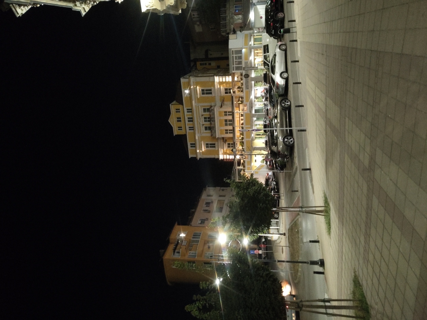 The image shows a street scene at night in Ruse, Bulgaria. The foreground features a tiled sidewalk with two trees. Beyond the sidewalk is a street with parked cars, including a silver SUV and a white van. In the background, there are several buildings illuminated at night. One prominent building is a yellow, multi-story structure with numerous windows and ornate architectural details. Another building to the left is orange and appears to be an apartment complex. Streetlights are visible, casting a glow on the scene. The sky is dark, indicating it is nighttime.
