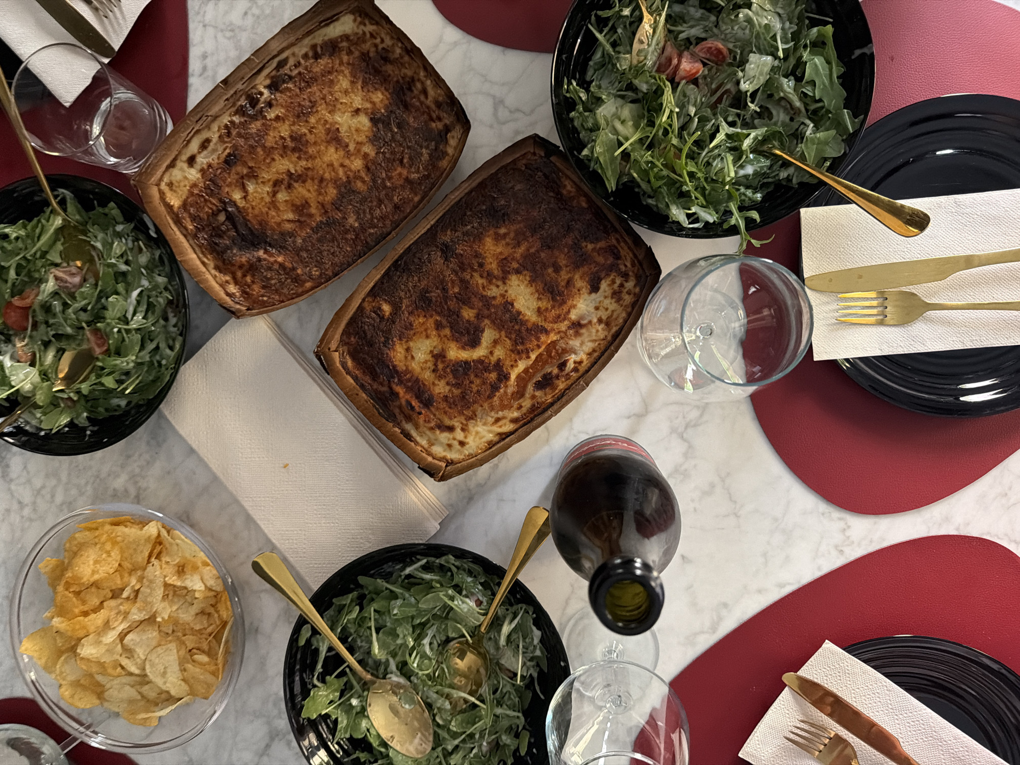 An overhead shot captures a dining table set for a meal in Milan, Italy. The table, with a white marble-like surface, displays a variety of dishes and place settings, suggesting a casual gathering or dinner party.
In the center of the table are two rectangular baked dishes, likely quiches or savory tarts, featuring a golden-brown, slightly charred crust. Surrounding these main dishes are several accompaniments. Three black bowls hold fresh green salads, primarily arugula, tossed with a creamy dressing and garnished with cherry tomatoes. Gold-colored spoons are placed in each salad bowl. A clear glass bowl is filled with a generous serving of potato chips.
The table settings include black dinner plates resting on unique, irregularly shaped red placemats. Each setting features a white folded paper napkin with elegant gold-colored cutlery – a knife and a fork. Various clear glassware is present, including stemless drinking glasses and wine glasses. A dark glass bottle, possibly wine, with a red label (text not discernible) and an open neck, is positioned near one of the wine glasses.
The scene depicts a prepared meal, awaiting guests or mid-service, creating an atmosphere of relaxed dining. No individuals are visible, and no specific time of day or weather cues are present beyond the indoor setting with artificial lighting. There is no clearly visible text on any of the objects.
