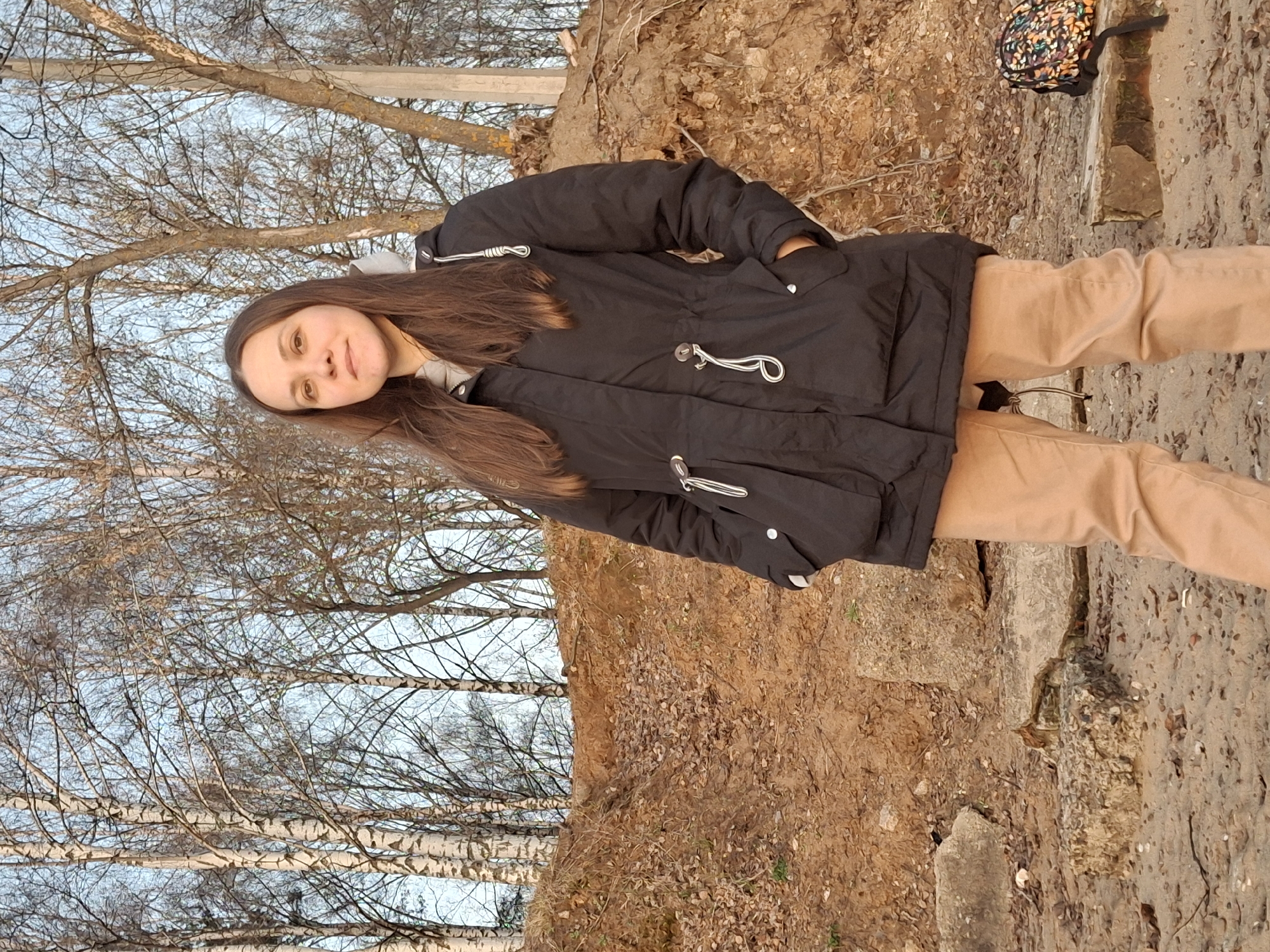 A medium shot shows a young woman in Chaikovsky, Russia, standing outdoors. She wears a black winter coat, beige trousers, and a grey top underneath the coat. Her hands are in her pockets. Her hair is long and brown. The background consists of bare birch trees and a slope of brown earth, transitioning to a cement surface near her feet. A colorful backpack rests on the cement surface to the upper right. The lighting suggests a late afternoon or early morning scene.