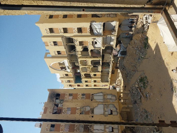 The image shows a severely damaged urban landscape in Khan Yunis, Palestinian Territory, under a clear blue sky. Two multi-story, beige-colored residential buildings dominate the scene, both exhibiting extensive structural damage. The building on the left features numerous small impact perforations across its facade, large sections of missing exterior walls exposing interior rooms, broken windows, and prominent black burn marks on its upper floors. Water tanks are visible on its rooftop. The building on the right, distinguished by decorative arched architectural features, also has significant portions of its exterior walls and balconies destroyed or missing, particularly across its lower and middle levels, revealing shattered interiors. White tarpaulins or fabrics are draped over exposed sections of both buildings.
The ground between and in front of the structures is sandy, heavily disturbed, and covered with substantial piles of concrete rubble, twisted metal, and general debris. Four adult males are observed standing together near the base of the right building, amidst the rubble. A single child sits on the sandy ground in the lower-left foreground. A white tent-like structure is partially visible in the lower-right foreground. The scene is partially framed by dark, vertical metal elements, suggesting the photograph was taken from an elevated position within another structure.