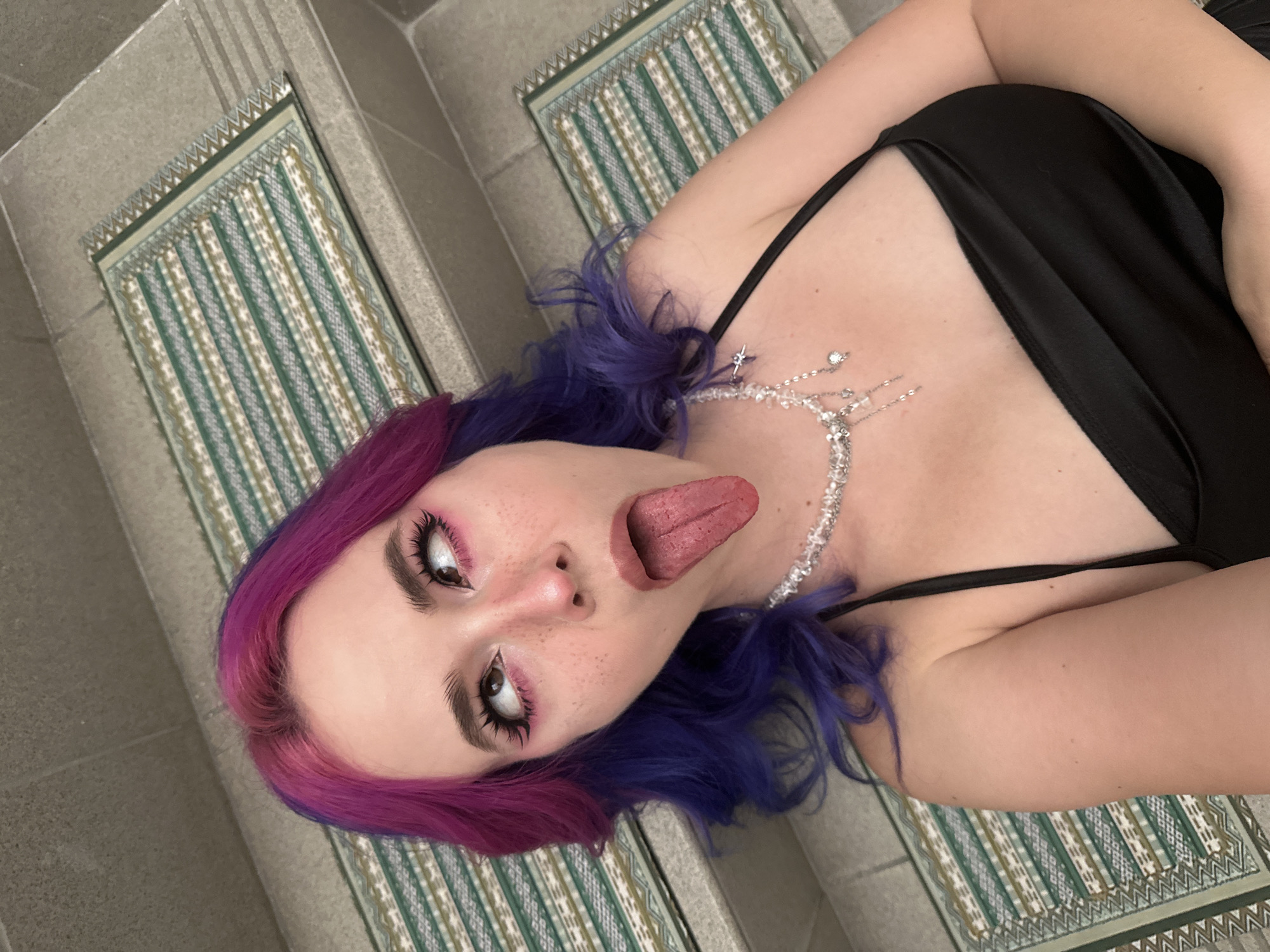 An overhead, close-up shot captures a person lying on their back, looking upwards with their tongue extended in a playful or provocative expression. The individual has vibrant two-toned hair, transitioning from pink at the roots to purple ends. Their face shows prominent eye makeup, including pink eyeshadow, dark winged eyeliner, and long, dark eyelashes complementing their well-defined eyebrows. Light freckles are visible across their nose and cheeks.
They are wearing a black top or dress with thin shoulder straps. A sparkling necklace, seemingly made of clear or silver-colored beads and small star-shaped elements, drapes across their chest. The background features light grey concrete or tiled flooring, which appears to be steps or a textured surface. Two rectangular decorative mats are inset into the floor, displaying a patterned design with vertical stripes and geometric borders in shades of green, white, and a light tan or gold color. There is no visible text in the image.