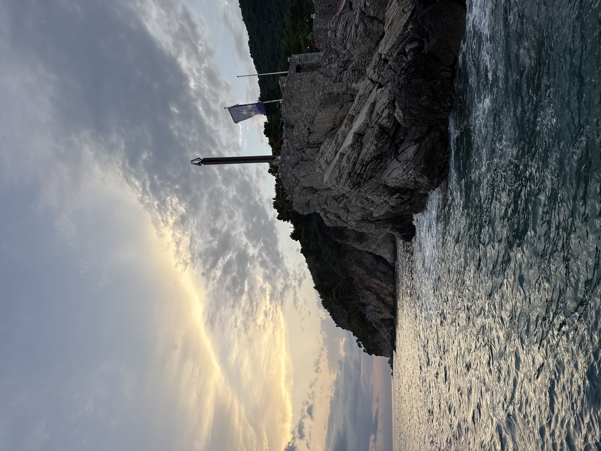 The image shows a rocky coastline with a stone structure built atop it. A tall, dark tower with an unusual, pointed top stands on the structure. A blue flag with yellow stars is also visible, flying from a pole. People are present on the structure, some standing and others seated. The sea is in the foreground, with waves crashing against the rocks. The sky is cloudy, with the sun setting and casting a warm glow on the clouds. The location is Petrovac centar, Montenegro. The scene is a picturesque coastal view at dusk, with a mix of natural and man-made elements.
