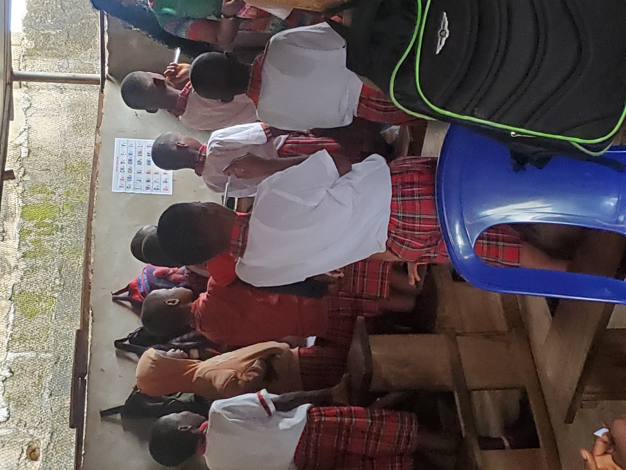 The image captures a classroom scene in Ukwu-Abwa, Nigeria. A group of children, likely primary school students, are seated at desks, dressed in a school uniform that includes a white shirt, red and plaid patterned skirt or shorts, and various colored long sleeve undershirts. A chart of the alphabet is affixed to the wall near the front of the classroom. One blue plastic chair and a backpack are visible. The classroom environment appears to be simple and somewhat cramped.