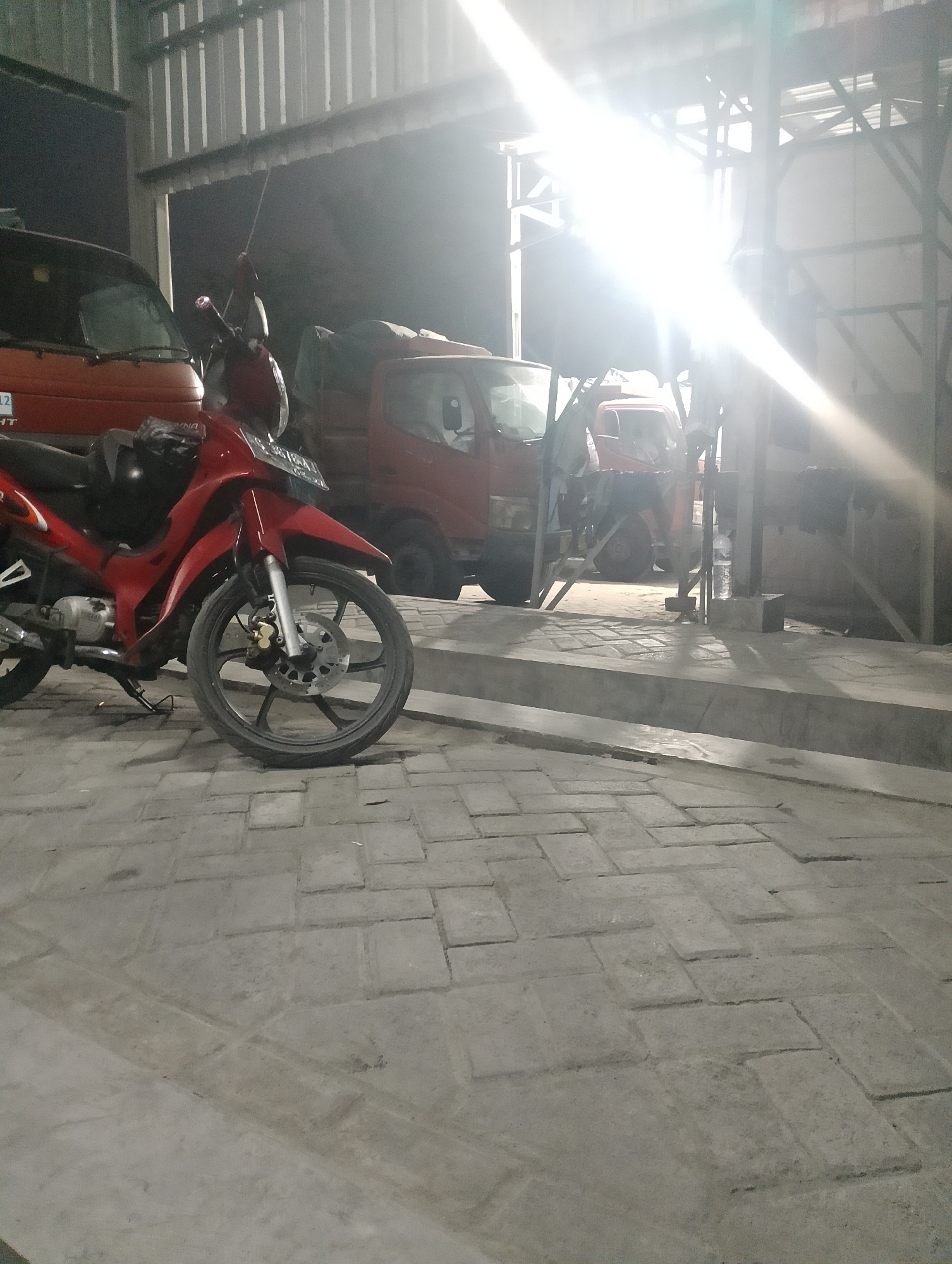The image depicts an outdoor scene at night, possibly within a covered area like a garage or workshop, in Jambean, Indonesia. In the foreground, a red motorcycle is parked, angled to the left. It has a black seat and a black helmet resting on it. The front wheel of the motorcycle is visible, with a disc brake. Behind the motorcycle, two red trucks are parked. The truck on the left is partially obscured by the motorcycle. The truck on the right is more fully visible, with its cabin and a covered cargo bed. The environment appears to be paved with rectangular stones, forming a walkway or yard. The background shows a metal roof structure and bright overhead lighting, which creates glare and washes out some details. The overall lighting suggests it is nighttime. There are no people visible in the image.