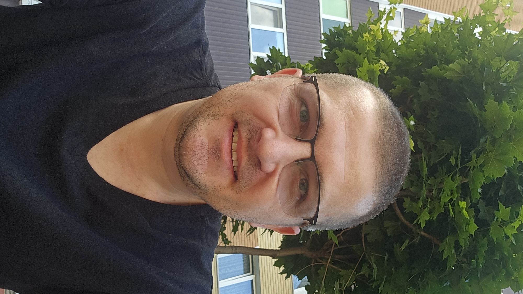 The image shows a man wearing glasses and a black V-neck t-shirt, posing for a selfie. He has short, dark hair and a light beard. Behind him, a building with horizontal brown and beige siding and white-framed windows is visible, along with lush green foliage from a tree. The scene appears to be outdoors, possibly on a sunny day, given the lighting. The building's architecture suggests an urban setting. Without further information or identifying markers within the image itself, it is impossible to definitively ascertain the specific location within Kyiv, Ukraine.