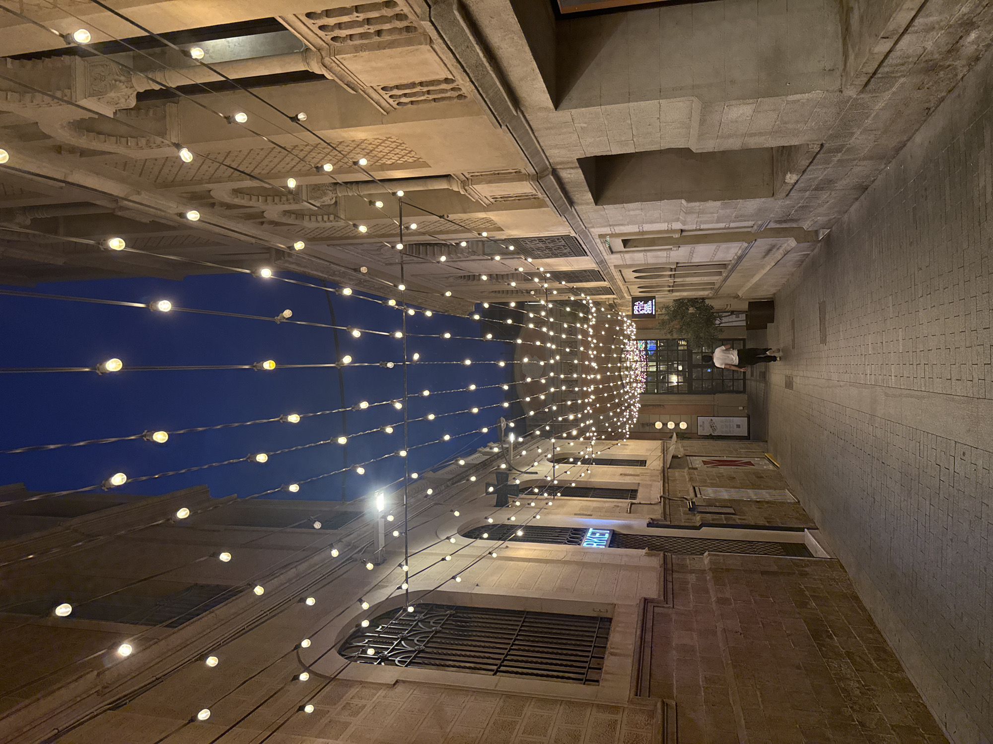 The image depicts a pedestrian alleyway at dusk, likely in the Gothic Quarter of Barcelona, Spain, illuminated by strings of festoon lights suspended overhead. The architecture on either side of the alley features aged stone facades, arched windows, and decorative details indicative of historic European city centers.
On the left, a building with a rust-colored stone lower section and lighter upper facade displays barred windows. A neon sign reading "MARKET" in a blue glow is visible on this side. The alley floor is paved with gray cobblestones.
On the right, a building with similar stone construction exhibits more elaborate architectural elements, including decorative carvings and balconies.
A lone individual, seen from behind, walks away from the camera down the alley. This person is wearing a white t-shirt and dark trousers. The sky above is a deep twilight blue, transitioning from lighter hues near the horizon. The overall atmosphere is tranquil and atmospheric, suggesting an evening stroll through a charming urban environment.