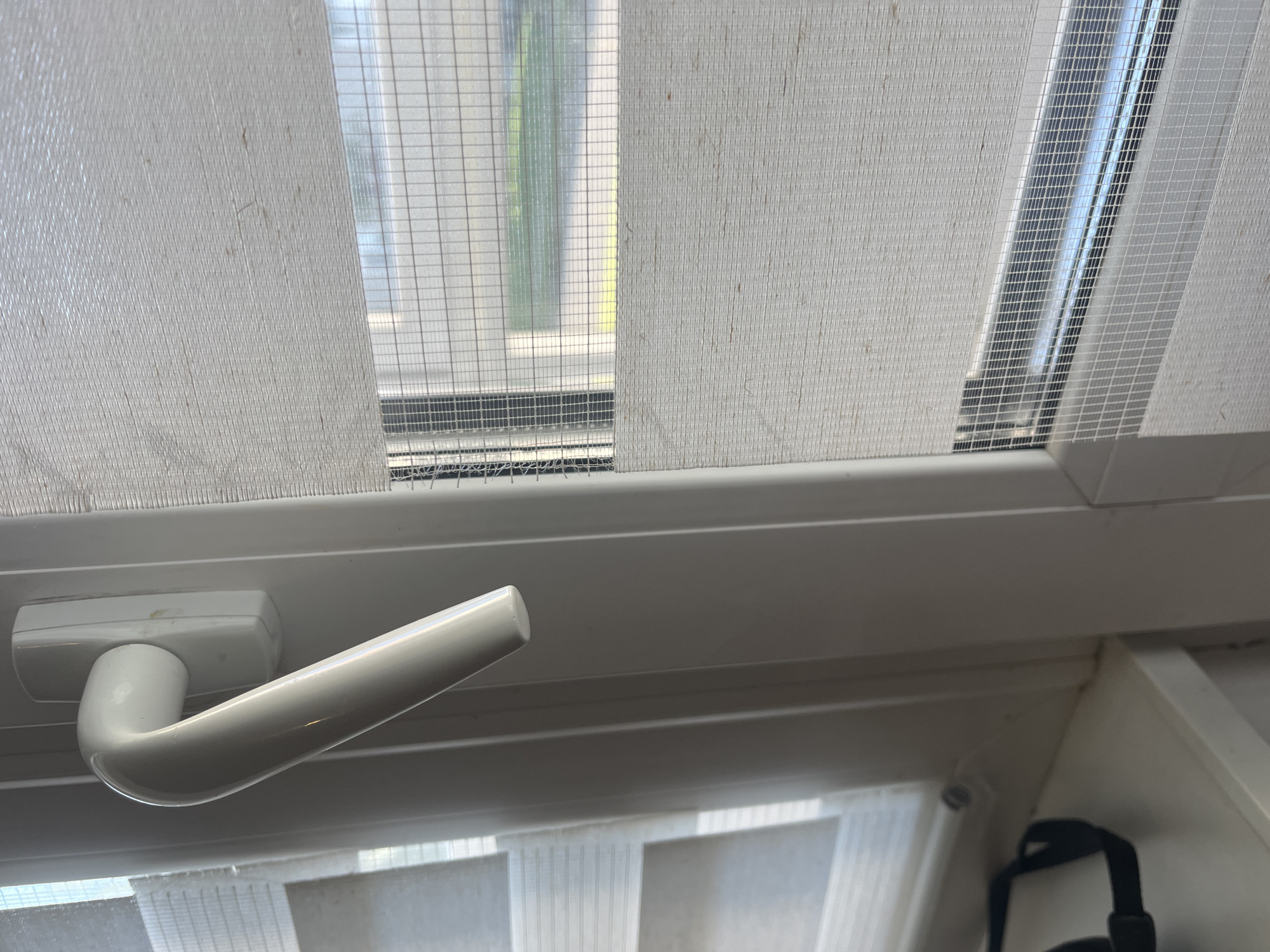 The image shows a close-up view of a white window frame with a white handle. The window is fitted with a day-night roller blind that alternates between opaque and sheer fabric panels. A fine mesh screen is visible behind the sheer panels, suggesting the window can be opened while keeping insects out. The scene is indoors, with sunlight filtering through the blinds, casting shadows and creating a bright, airy atmosphere. The window appears to overlook a blurred outdoor scene with green foliage and a building. Part of a black strap, possibly from a bag or camera, is visible on the white windowsill in the lower left corner. The overall impression is of a domestic or office setting. The image does not contain any elements that would allow for pinpointing its location within the city of Lahojsk, Belarus.