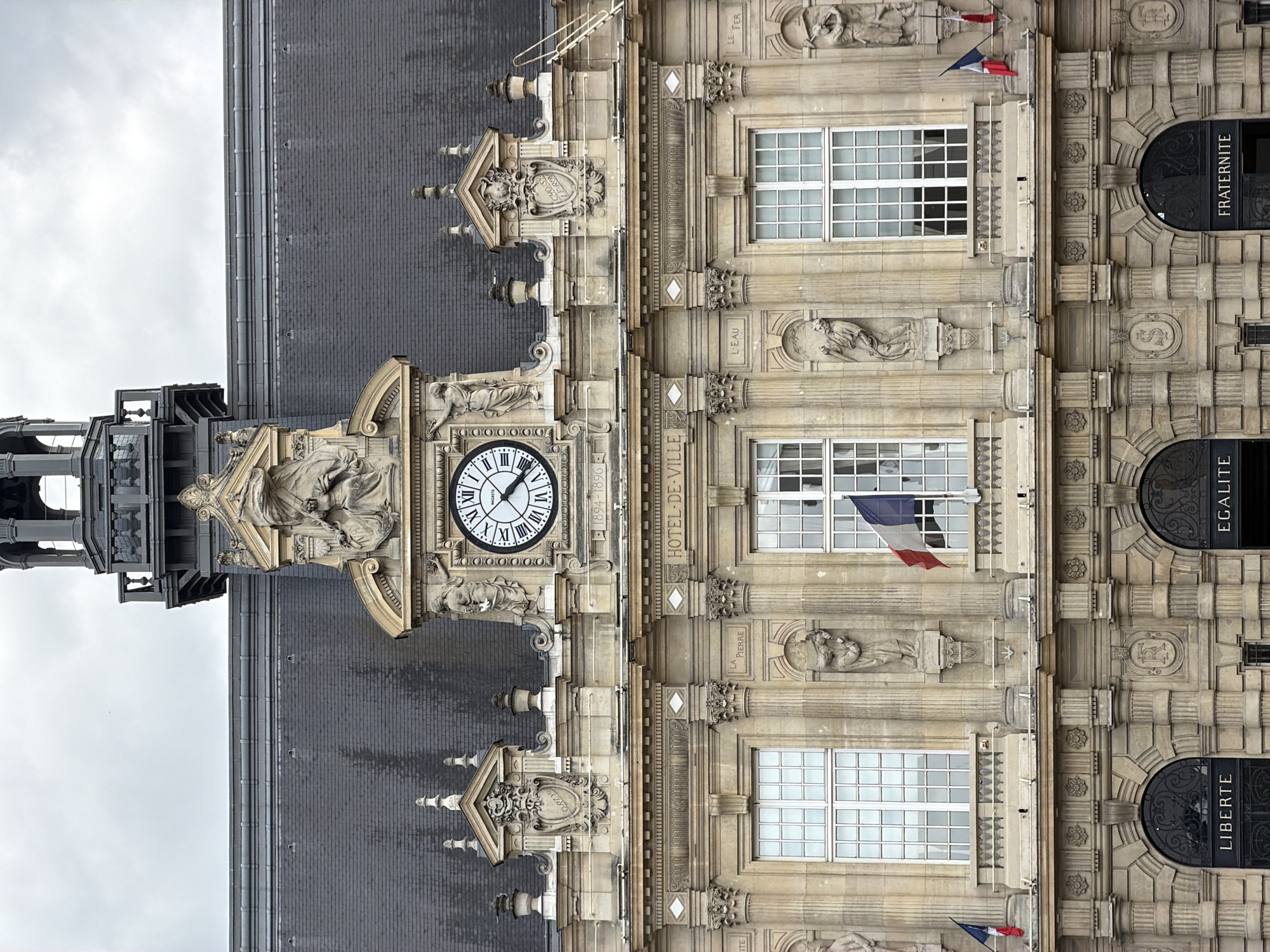This media file showcases a detailed, vertical, exterior view of a grand stone building, likely a historic public edifice. The architecture is ornate, featuring classical columns, intricate sculptures, and decorative stonework. A prominent clock face with Roman numerals is centrally located, indicating the time. French flags are visible, suggesting the location is in France.
The building's facade is divided into sections, with arched doorways bearing the inscriptions "LIBERTE", "EGALITE", and "FRATERNITE". The building's name, "HOTEL-DE-VILLE", is also visible, confirming it is a town hall. The dates "1894-1896" are inscribed, likely indicating the construction period.
The weather appears overcast, with a bright, diffused light. The sky is cloudy, creating a muted atmosphere. Sculptural figures are integrated into the facade, depicting allegorical or historical subjects. The overall impression is one of architectural grandeur and civic importance. The style of the building, combined with the French motto, strongly points to a location within France, and the OCR confirms it is the Hotel-de-Ville in Ivry-sur-Seine.
---
The provided OCR text includes "CHOISY LE-ROV", which is a different town than Ivry-sur-Seine, and also "ORZY" and "VAY" which are not immediately identifiable as locations in this context. Given the prominent "HOTEL-DE-VILLE", "LIBERTE", "EGALITE", "FRATERNITE", and the architectural style, it is highly likely a French town hall. The presence of "1894-1896" suggests a historical building. The image is a close-up, emphasizing the architectural details and the clock.