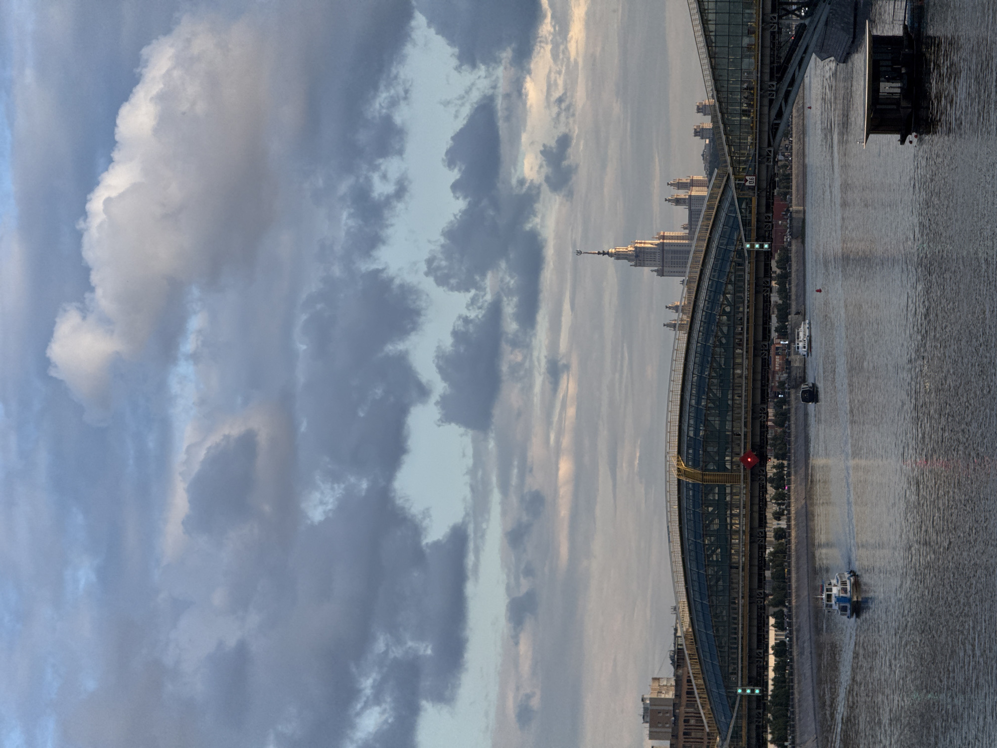 The image depicts a view of the Moskva River in Moscow, Russia, under a cloudy sky with patches of blue. A modern, arched bridge with glass and metal structures spans the river, with a prominent skyscraper in the background, likely the Moscow State University building. Two boats are visible on the river: one is a white and blue passenger boat moving away from the viewer, and another is a smaller, dark vessel farther downriver. A small, dark, wooden structure resembling a boathouse or floating kiosk is also present on the water. The riverbanks are lined with trees and buildings. The lighting suggests it is late afternoon or early evening.