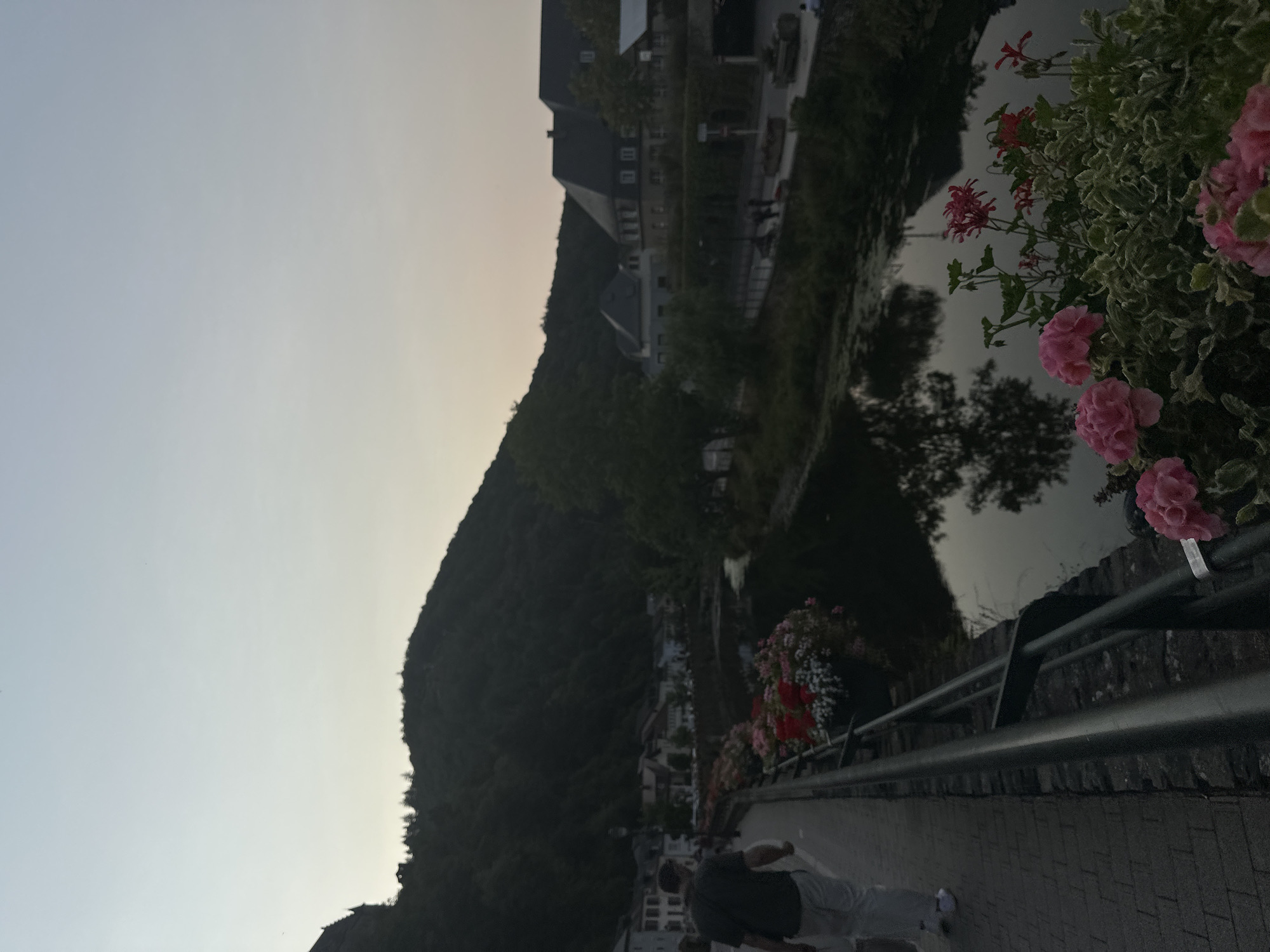 The image shows a man walking on a paved walkway alongside a river in Vianden, Luxembourg. The walkway is lined with a metal railing and planters filled with pink and red flowers. In the background, there are buildings with traditional European architecture, and a dense, tree-covered hill rises behind the town. The sky is overcast and the light suggests it is either early morning or late evening. The river is calm and reflects the surrounding environment. Several other people are visible in the distance walking on a street across the river.