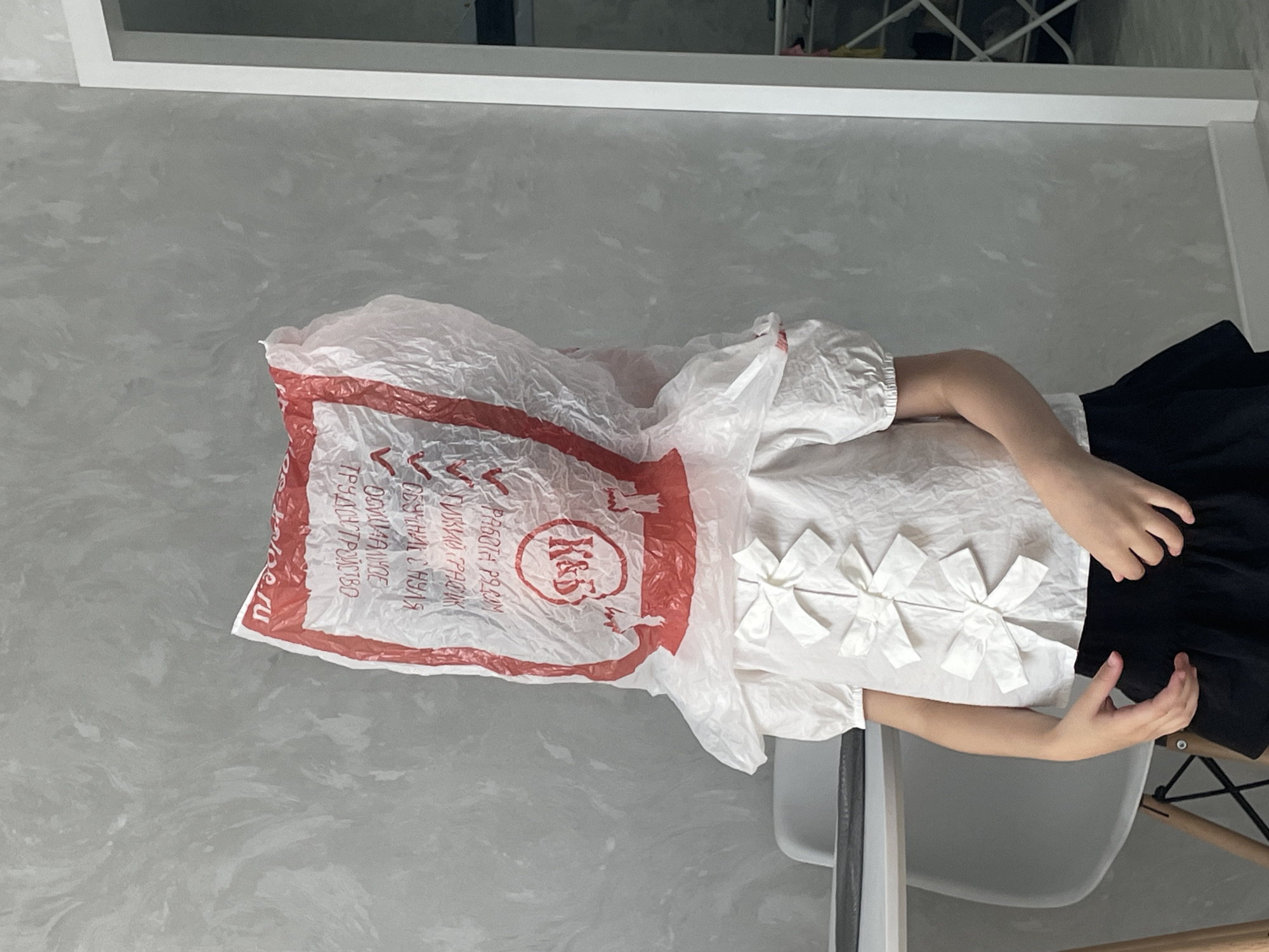 The image captures an indoor scene, likely a home in Izhevsk, Russia, showing a person with a white plastic bag covering their head. The individual is seated at what appears to be a table, partially visible on the right, resting their arms on its surface. They are wearing a short-sleeved white blouse adorned with several decorative white bows down the front, paired with a dark (likely black) skirt or trousers. Their hands are visible, one resting on the table and the other casually placed on their lap/skirt.
The white plastic bag has prominent red text and a logo. The logo, enclosed in a red circle, reads "K&S". Other visible Cyrillic text in red includes "РАБОТА РЯДОМ" (Work nearby), "ОБУЧИМ С НУЛЯ" (We will train from scratch), "ТРУДОУСТРОЙСТВО" (Employment), "ОФИЦИАЛЬНОЕ" (Official), and part of a web address ".ru" (possibly "pole.ru" or similar). This text suggests the bag might be from a business related to job placement or training.
The background features a light gray wall with a subtle, swirling marble-like pattern. Above this, a white horizontal shelf or frame is visible, with glimpses of a darker area or shelving unit behind it. The lighting is uniform, suggesting an indoor setting during the daytime or under artificial lights. The person's posture appears relaxed, and the act of wearing a bag over the head could be playful or an attempt to obscure identity.