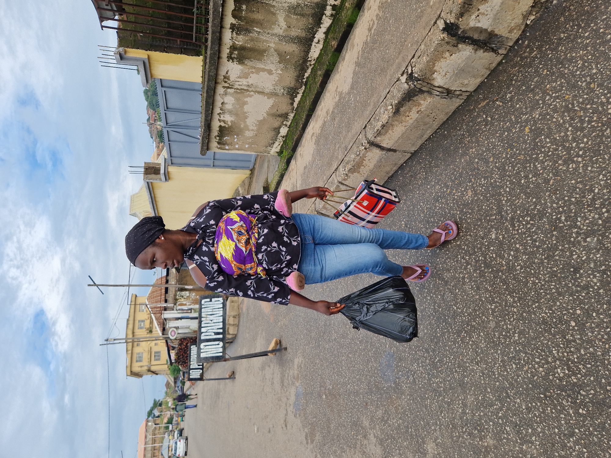A medium shot captures a young Black woman from the knees up walking on an asphalt road in Kabong, Nigeria, on a cloudy day. She is wearing blue jeans, pink flip-flops with blue painted toenails, and a black off-the-shoulder shirt with a colorful patterned fabric across the chest. A pink baby carrier with a baby is strapped to her front. She carries a black plastic bag in her left hand and a red, white, and blue plaid tote bag in her right. Two "NO PARKING" signs are visible behind her to the left. A building with yellow walls and a gray metal gate stands to the right, with a cracked concrete wall in the foreground. The road is bordered by a concrete curb.