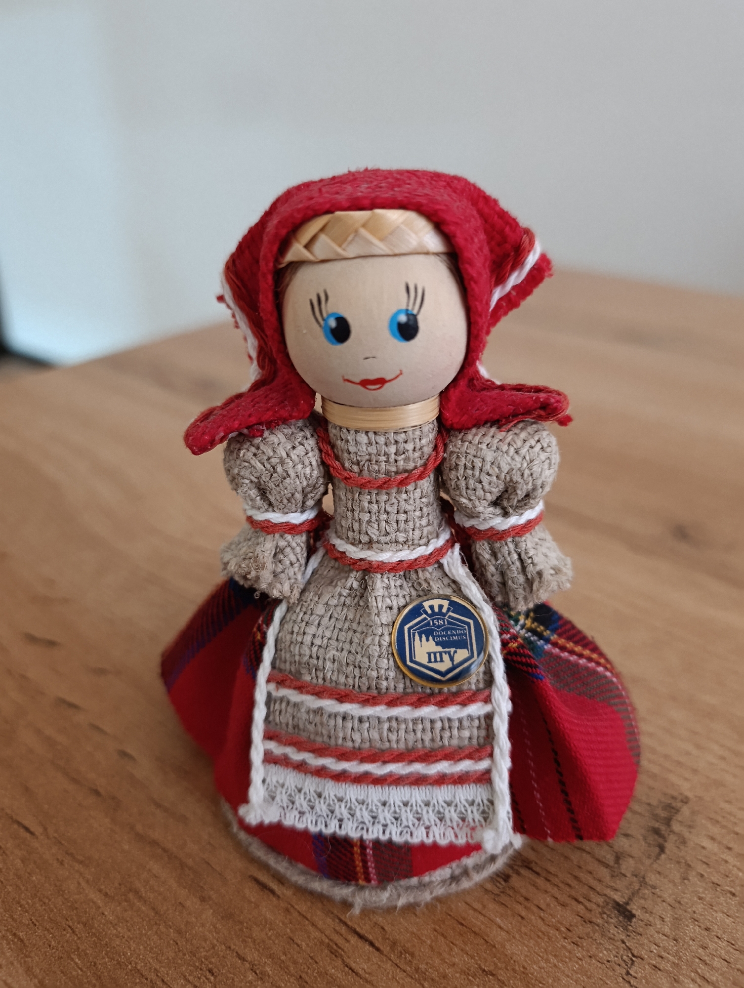 A close-up, eye-level, full shot shows a small, fabric doll dressed in traditional clothing, resting on a wooden surface. The doll has a round, wooden head with painted blue eyes, eyelashes, and red lips. It wears a red headscarf with white trim, a rough, beige fabric bodice with puffed sleeves, and a layered skirt consisting of a beige apron over a red and plaid fabric underskirt. Red and white trim accent the doll's dress. A circular blue and gold pin with text and an image is attached to the front of the doll's apron. The wooden surface appears to be a table with a visible grain. The background is out of focus, showing a light-colored wall. The doll does not appear to be located in the city of Termez, Uzbekistan, as there are no discernible landmarks or context to suggest this location.
