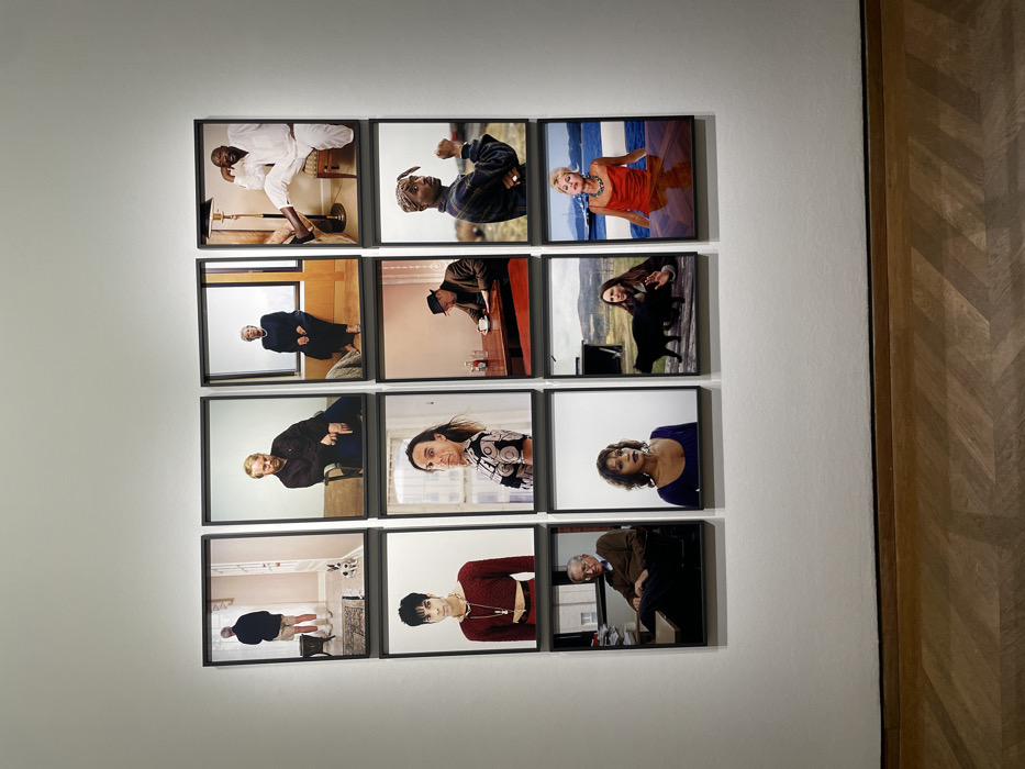 Twelve framed portraits are hung in a grid formation on a white wall. The portraits are diverse, featuring individuals of varying ages, ethnicities, and genders. Some subjects are dressed casually, while others wear more formal attire. The photographs have different indoor or outdoor backgrounds. Each is framed in a simple black border. A strip of wood paneling is seen on the right side of the frame.