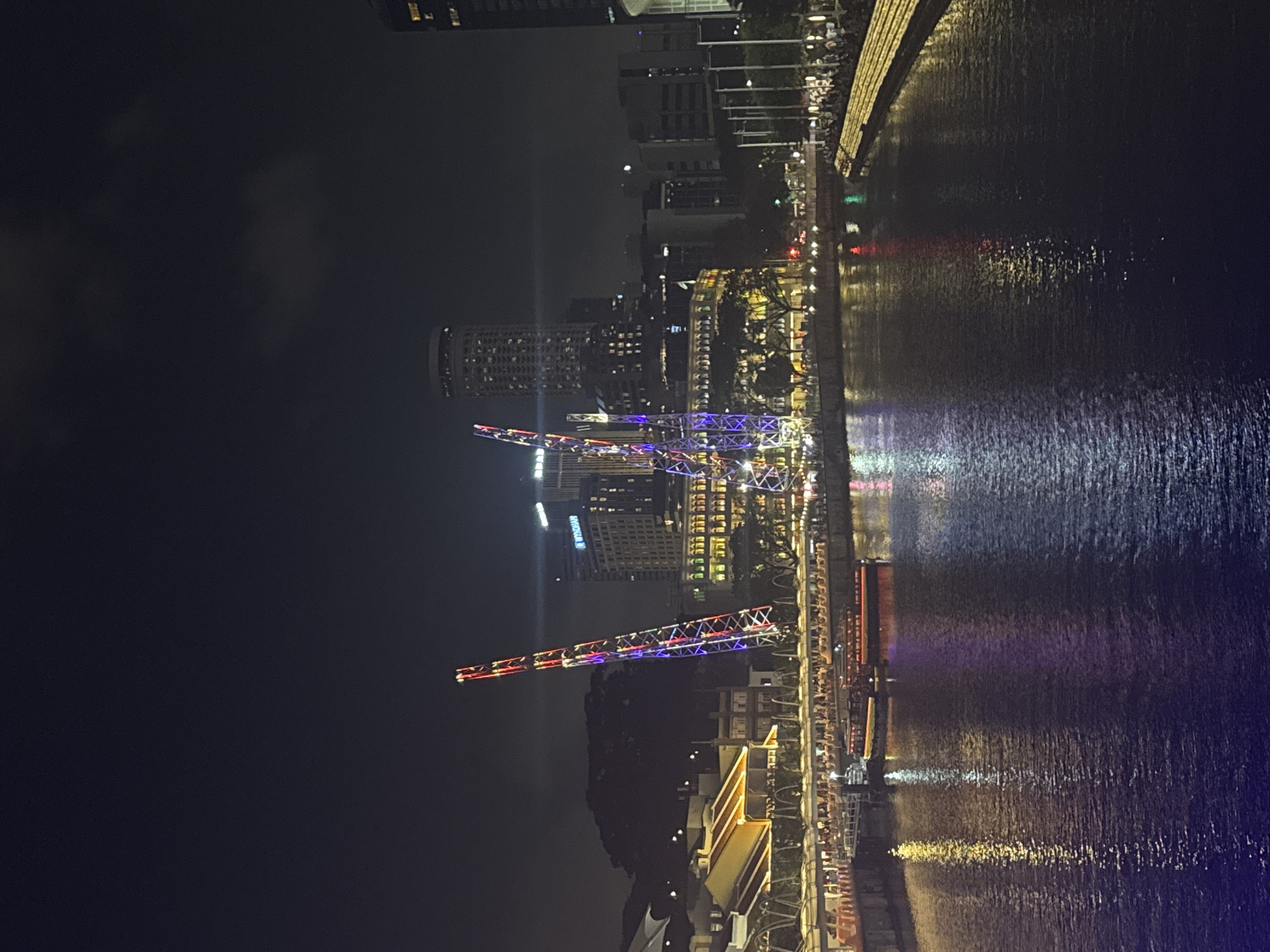 This is a nighttime photograph of the Singapore River at Clarke Quay. Two tall, brightly lit structures, resembling amusement park rides or decorative towers, dominate the left side of the frame, displaying red, blue, and white lights. In the background, various buildings are illuminated, including a circular tower with many windows and a tall, modern skyscraper with the "WYNDHAM" logo visible.
A body of water, presumably the Singapore River, occupies the foreground and midground, reflecting the city lights. Several boats are docked or moving on the river, including a longer, red and yellow passenger boat. Along the riverbanks, there are illuminated walkways and a dense cluster of people gathered, particularly on the right side where a tiered, illuminated structure is visible. The scene depicts a vibrant urban waterfront area at night.