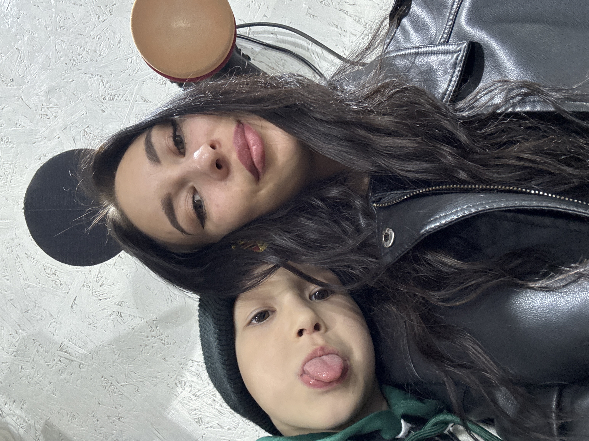 This portrait captures two individuals, likely a mother and child, in a close-up, angled selfie. The adult female, positioned higher in the frame, has long, dark, wavy hair, distinct dark eyebrows, and is wearing makeup including dark eyeliner and pink lipstick. She looks directly at the camera with a soft smile and is clad in a black leather jacket.
Below her, a young child also faces the camera, playfully sticking out their tongue, conveying a mischievous and lighthearted expression. The child is wearing a dark beanie hat and a green hoodie.
The background consists of a white, textured wall, possibly painted oriented strand board (OSB) or a similar material with visible wood chip patterns. Partially visible at the top left are elements resembling Mickey Mouse ears – a black circular shape and a tan, rounded object, suggesting a novelty headband or decorative item. The overall scene depicts a casual, affectionate, and playful indoor moment between the two subjects, potentially in Chaikovsky, Russia. There is no visible text in the image.