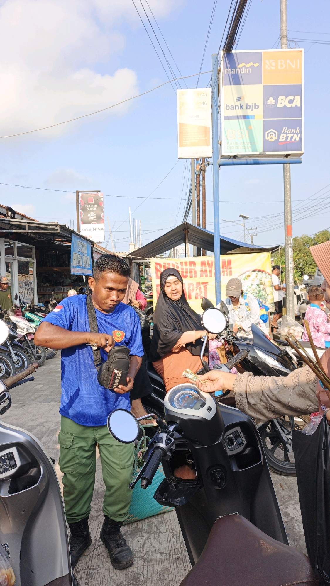 In Cibenda, Indonesia, on a sunlit street, a man in a blue t-shirt and green cargo pants is seen standing next to a motorcycle, rummaging through a small black pouch. He is wearing black boots and has a lanyard around his neck. To his right, a woman in a black hijab and orange top is seated on another motorcycle, looking towards the man. She is holding money in her hand, suggesting a transaction. Other individuals are present in the background, including a person in a pink shirt and another in a light-colored jacket. Several motorcycles are parked along the street, and stalls with signage are visible. The environment appears to be a bustling street market or commercial area, with utility poles and wires overhead. A large yellow banner with text in Indonesian is displayed on a stall behind the woman. The overall scene depicts a casual interaction, possibly a purchase, taking place in an outdoor urban setting.