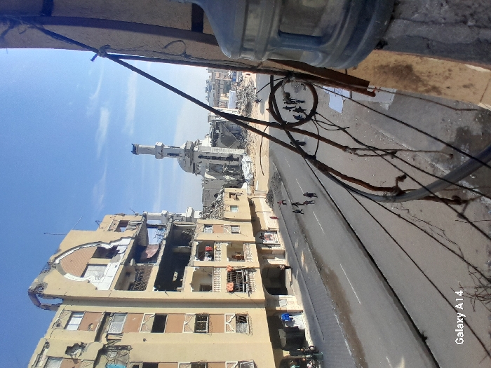 The image depicts an urban street in Khan Yunis, Palestinian Territory, characterized by extensive structural damage under a clear blue sky.
In the foreground, from an elevated perspective, the right side is partially obscured by a large, clear plastic water bottle and a cluster of dark, coiled wires and cables, suggesting the vantage point is a building's window or balcony.
Dominating the left midground is a multi-story residential building, light tan in color, with significant structural compromise. Multiple floors exhibit large sections of missing walls, exposing interior rooms and structural elements. Balconies on the lower levels are visible, some with laundry hanging.
An asphalt road runs horizontally across the midground, featuring a visible center median. The road surface appears uneven and possibly wet or stained in areas. Along the road and pavement, several small groups of people are walking; one group of approximately 6-8 individuals, including children, is visible in the right-center midground, moving away from the camera.
In the background, a large mosque with a tall minaret and a dome structure stands prominently, also heavily damaged. The dome shows partial collapse, and sections of the minaret appear broken or missing. Surrounding the mosque are extensive piles of rubble and debris, indicative of widespread destruction. Further into the distance, more damaged buildings and demolition sites are visible under the clear sky.