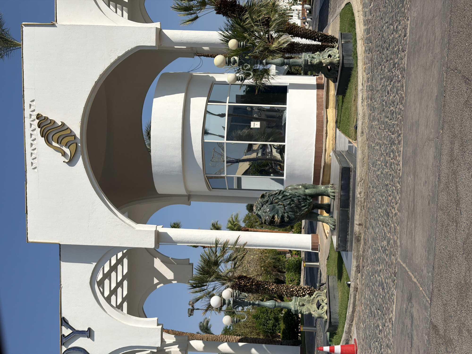 The image depicts an entranceway in Sharm El Sheikh, Egypt, likely to a resort or hotel, with a prominent white arched structure. The architecture features Arabic-inspired arches and a decorative seashell motif with golden accents at the top. In the foreground, a dark green, weathered bronze statue of a lion stands on a raised, dark tiled platform. To the left of the lion, a vintage-style lamppost with three globe lights is visible, also resting on a dark tiled platform. Another similar lamppost is positioned to the right of the lion. Behind the lion and lampposts, a circular white guardhouse with large glass windows reflects the surrounding palm trees and sky. The ground in front of the entrance is paved with cobblestones. A red and white traffic cone is placed on the left side of the cobblestone area. Palm trees and lush greenery are visible in the background. The sky is clear and blue, indicating a sunny day.