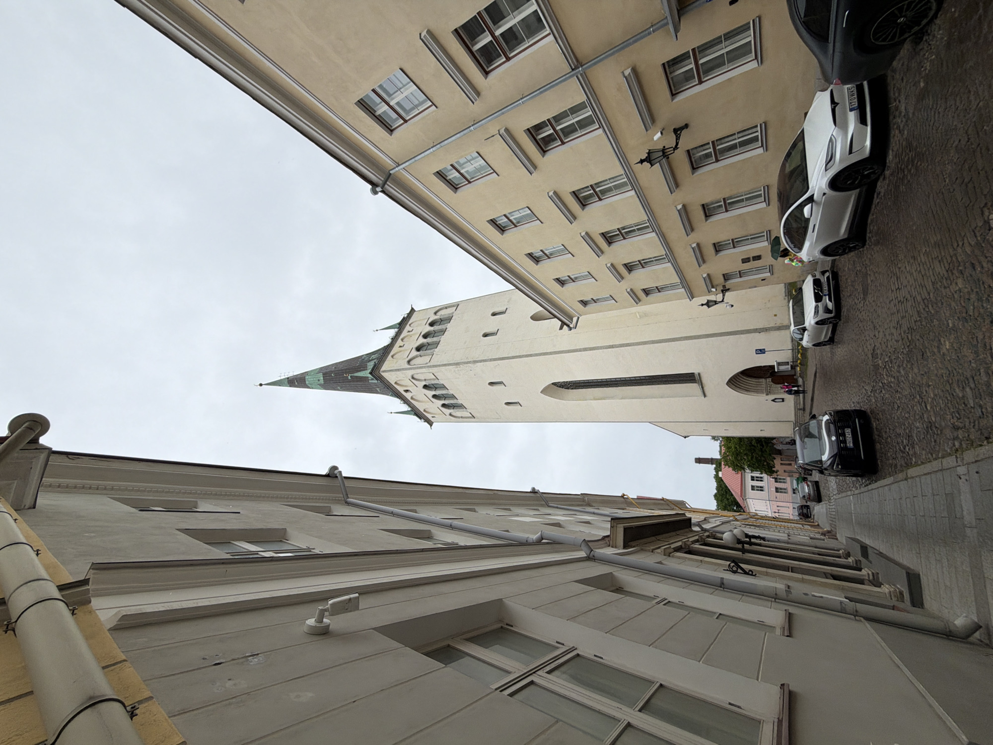 In Tallinn, Estonia, a view looking up a narrow, cobblestone street showcases historical architecture under a cloudy sky. Tall, pale buildings with cream and yellow facades line the street, featuring numerous windows with dark frames. A prominent white church tower with a green spire dominates the background, its gothic architectural style evident in the arched windows near the top. A few cars are parked on the street, including a black SUV, a white Tesla Model X, and a white Volvo station wagon. The street is paved with uneven cobblestones, and several lampposts are visible attached to the buildings. The scene is quiet, with no people clearly interacting. The perspective is angled upwards, emphasizing the height of the buildings and the church.