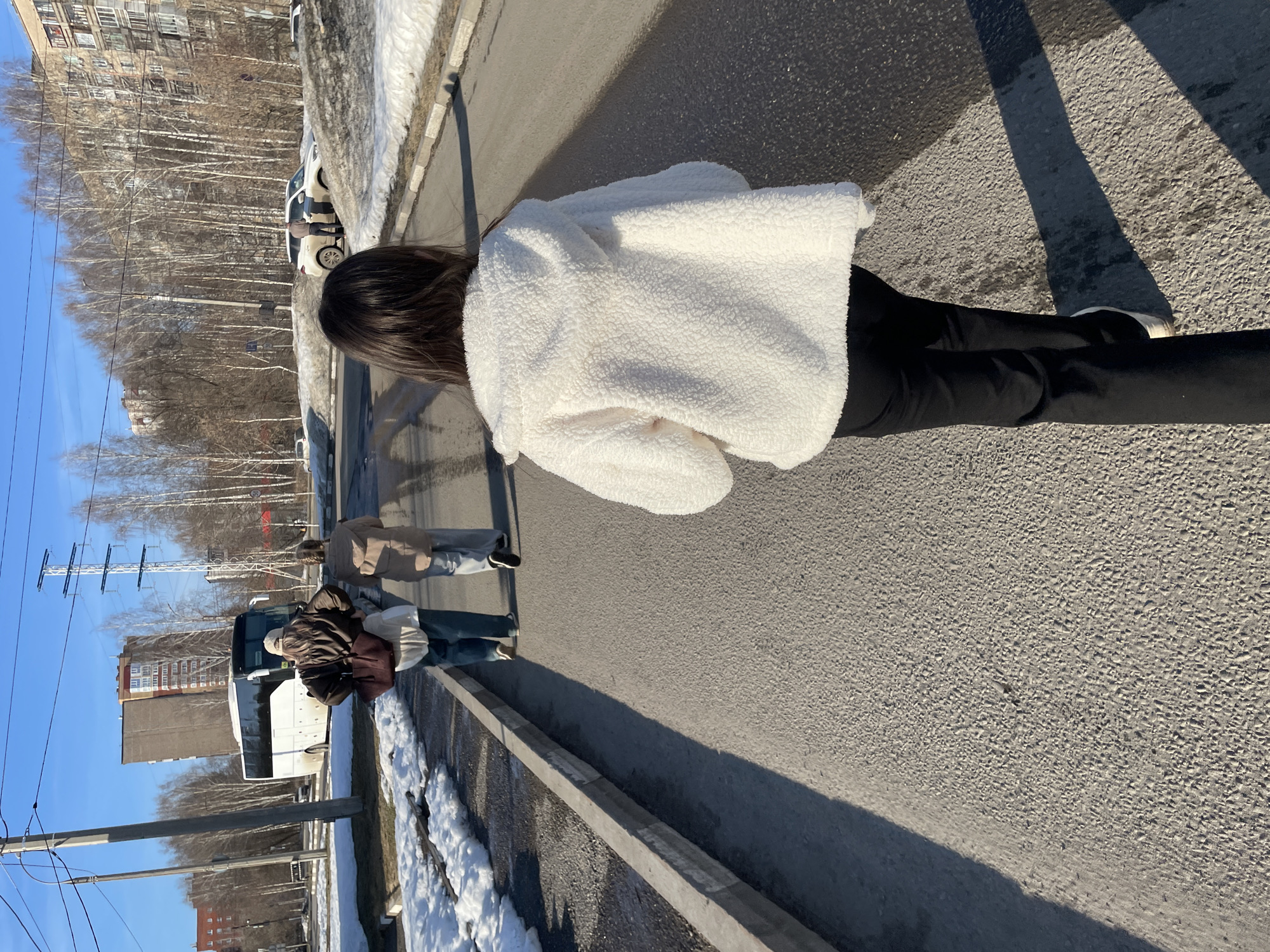 The image shows a person wearing a white, sherpa-like jacket and black pants walking on a paved road in what appears to be an urban area. Other people can be seen in the background walking near a white bus. Buildings and trees are visible in the distance, and patches of snow are along the roadside. Electrical power lines are visible in the background. The photograph was taken in Izhevsk, Russia.
