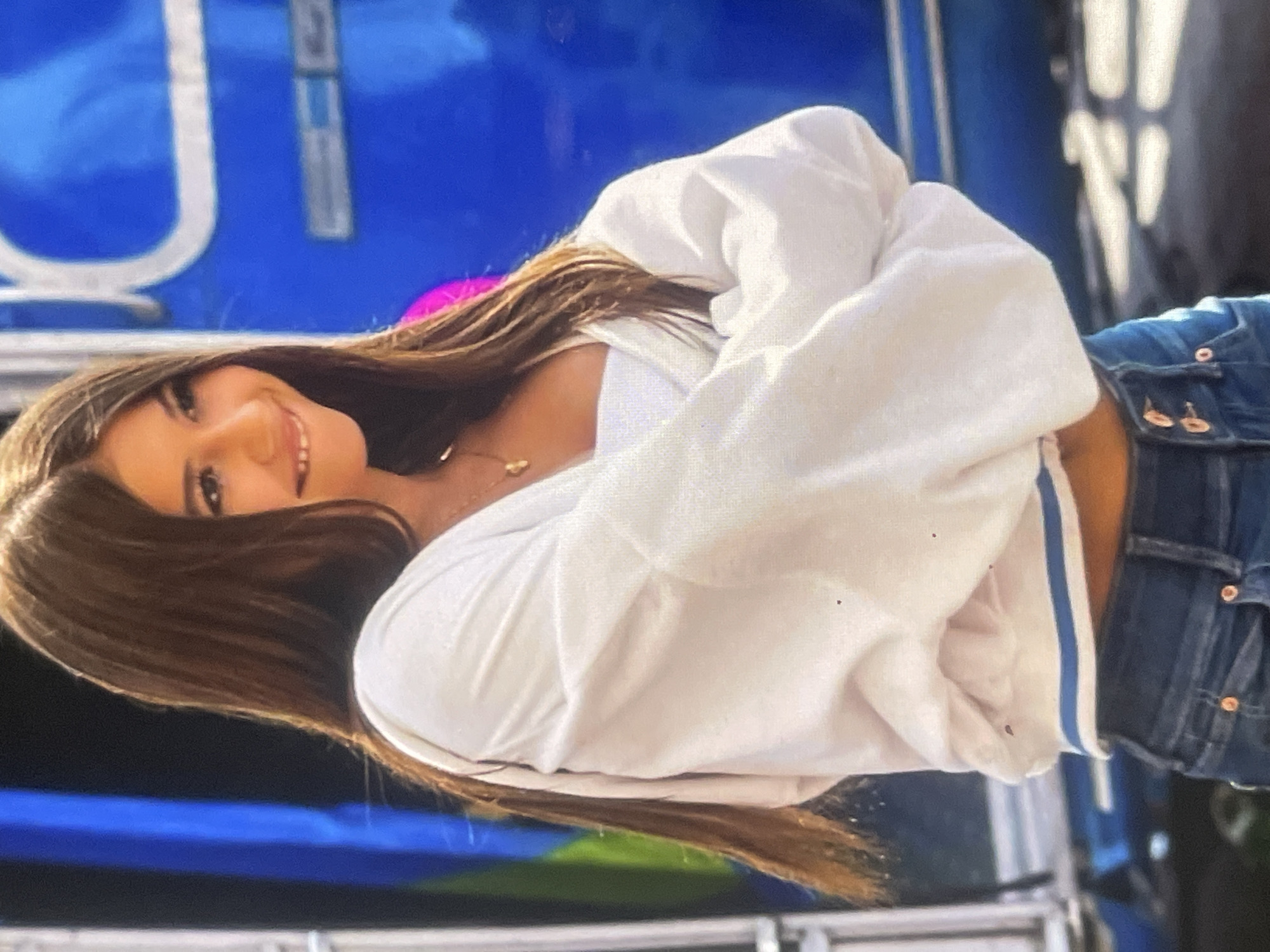 The image shows a young woman posing outdoors in what appears to be Poltava, Ukraine. She has long, straight dark brown hair, which falls over her shoulders, and is smiling broadly while looking directly at the camera. Her arms are crossed in front of her. She is wearing a white, long-sleeved cropped top or sweatshirt, revealing a sliver of her midriff, and blue denim jeans. A delicate gold necklace with small charms is visible around her neck.
The background features a large, bright blue banner or sign with prominent white lettering. Part of a large capital 'U' and what appears to be a 'T' or 'J' are discernible on the blue surface. A vibrant pink or magenta light source is visible behind her hair, possibly an artificial light or reflection. The lower portion of the background shows darker tones, suggesting shadows or other structures. The bright, clear lighting indicates it is daytime. The woman's relaxed pose and genuine smile convey a feeling of happiness and ease.