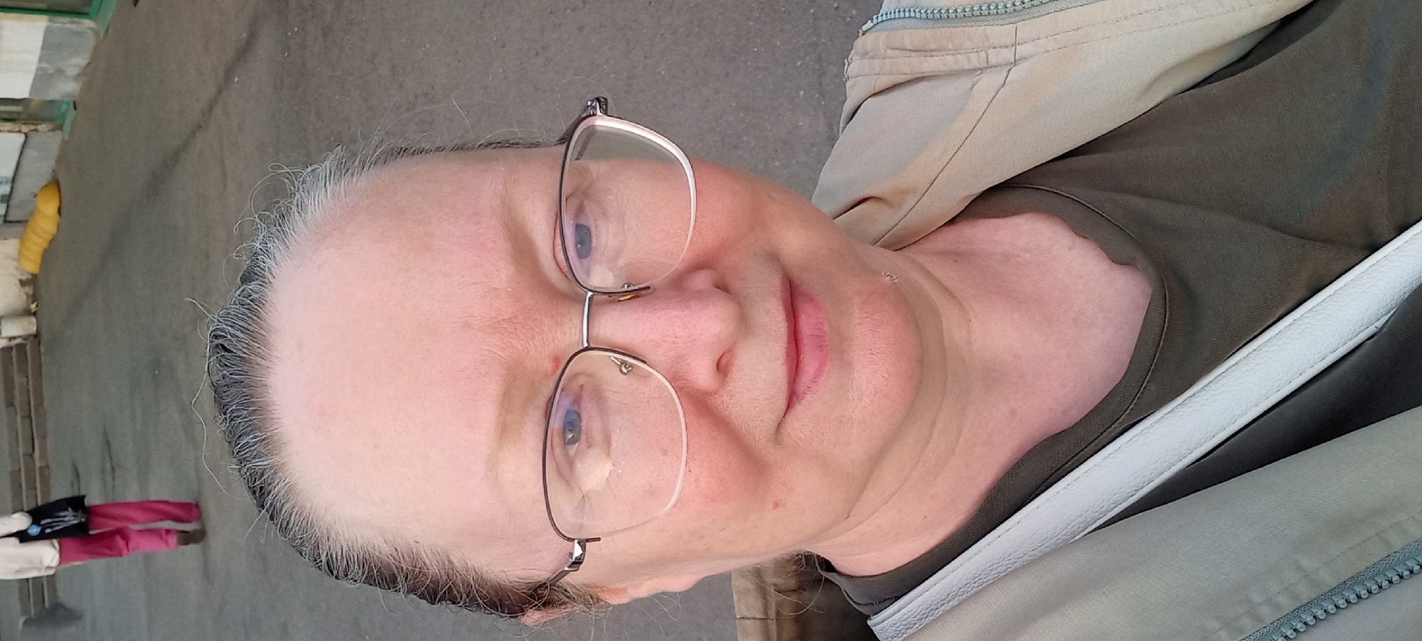 A middle-aged woman with graying hair and glasses is taking a selfie outdoors. She is wearing a dark green t-shirt and a light beige jacket. A white strap from a bag is visible across her chest. The background shows a grey paved area with some concrete stairs and a yellow cylindrical object, possibly a barrier, in the distance. The overall scene suggests a public outdoor space, such as a street or park.