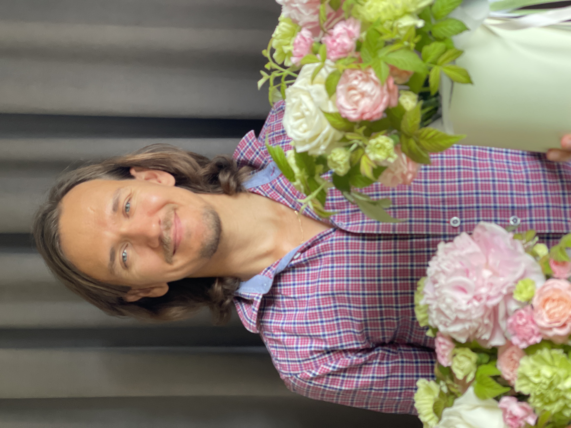 The image is a portrait of a man with long, brown hair and a beard, smiling at the camera. He is wearing a pink and blue plaid shirt. He is holding a bouquet of flowers, which includes pink and white roses, and green foliage. The background appears to be a gray curtain. The lighting suggests it could be daytime, but there are no definitive cues. The man's expression is happy and relaxed.