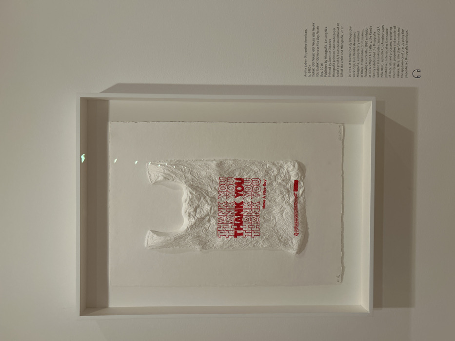 The image displays an artwork mounted on an off-white wall within a gallery setting, specifically identified as UCLA's Wight Art Gallery in Los Angeles, California, United States of America.
The central subject is a framed artwork titled "THANK YOU THANK YOU THANK YOU THANK YOU Have a Nice Day Plastic Bag," created in 2016 by Analia Saban (Argentine American, b. 1980). The artwork is a mixografia print on handmade paper, presented as an artist's proof (6/6). It depicts a highly textured, crumpled white form resembling a common plastic shopping bag, with multiple instances of the phrase "THANK YOU" printed in red capital letters across its surface, followed by "Have A Nice Day" in smaller red text. The print includes subtle red markings and text at the bottom edge, characteristic of store branding. The artwork is housed within a deep, white shadow box frame with a glass front, which reflects minor glints of light from an unseen source above.
To the right of the framed artwork, a descriptive text plaque provides details about the piece, the artist, and the printing technique, including the publisher Mixografia in Los Angeles. Below the text, a headphone icon indicates an audio guide is available. The environment is sterile and well-lit, typical of an art exhibition space, with no individuals visible in the immediate scene.