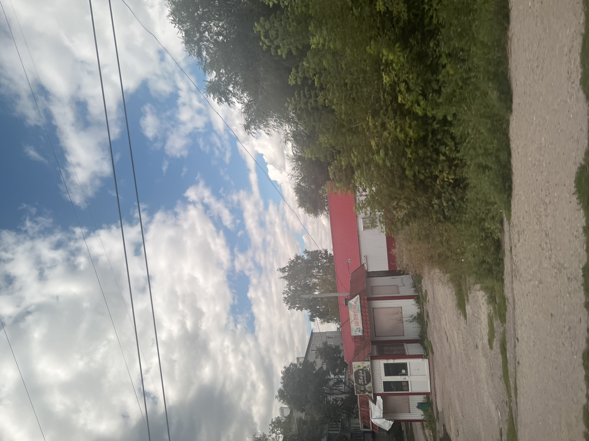The image shows a small, detached building with a red roof that appears to be a commercial establishment, possibly a cafe or a small restaurant, in Ulyanovsk, Russia. The building has white walls with red trim. Above the entrance, there are two signs. One sign, in Russian, reads "Кафе" (Cafe). Another sign displays an image of a shawarma and the word "ШАУРМА" (Shawarma) in Russian. The building is situated on an unpaved lot with some overgrown vegetation. To the left of the building, there is a multi-story residential building. To the right and behind the commercial building, there are dense trees and foliage. The sky is cloudy with patches of blue. Power lines are visible in the upper portion of the image. The scene appears to be an outdoor shot taken during the daytime.