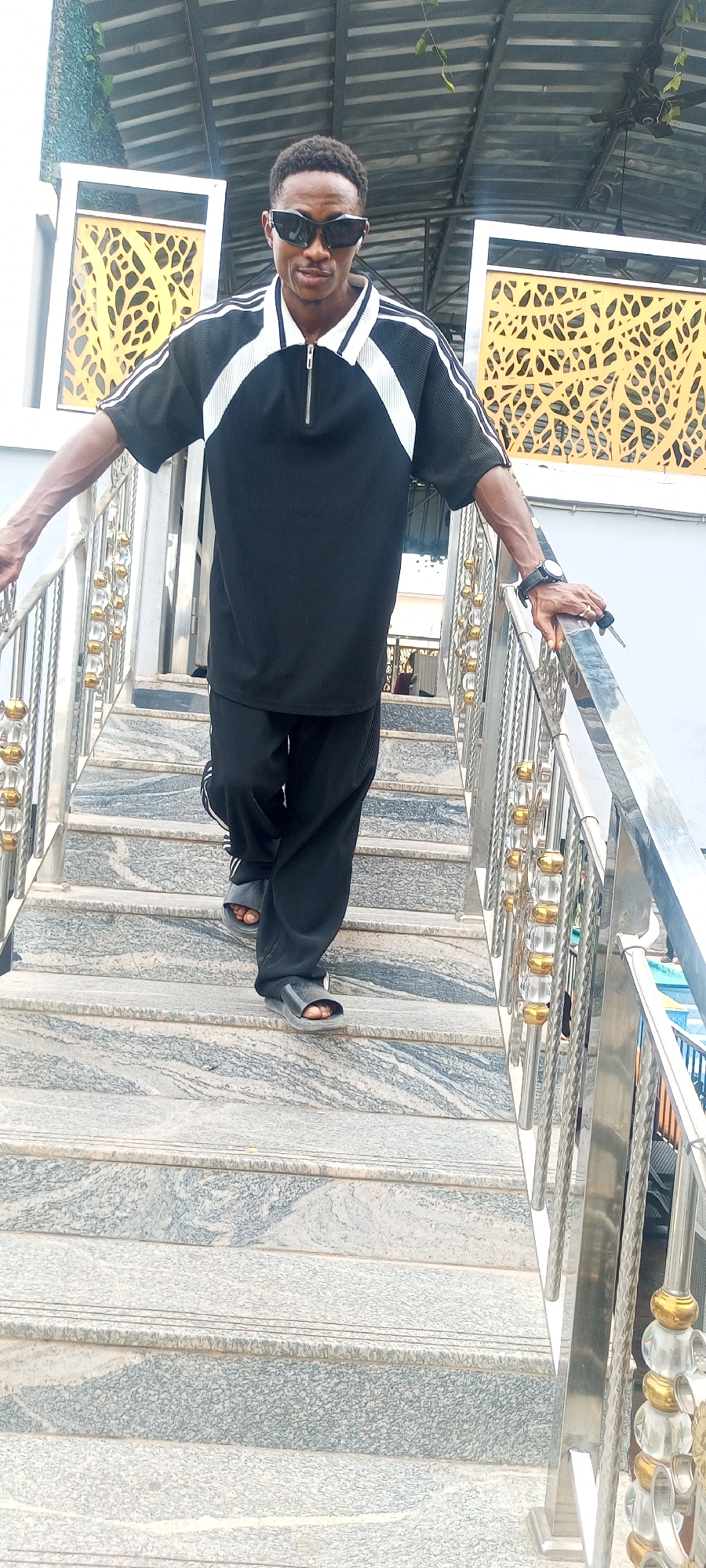 A man wearing a black athletic suit with white stripes and black sunglasses is walking down a set of outdoor steps. He is wearing black sandals and has a watch on his left wrist. He is holding onto the silver railing of the staircase with his left hand. The steps are made of patterned grey stone. The location appears to be outdoors under a partially covered structure. The city of Lokoja, Nigeria, is inferred, but there are no specific visual cues in the image to confirm this.