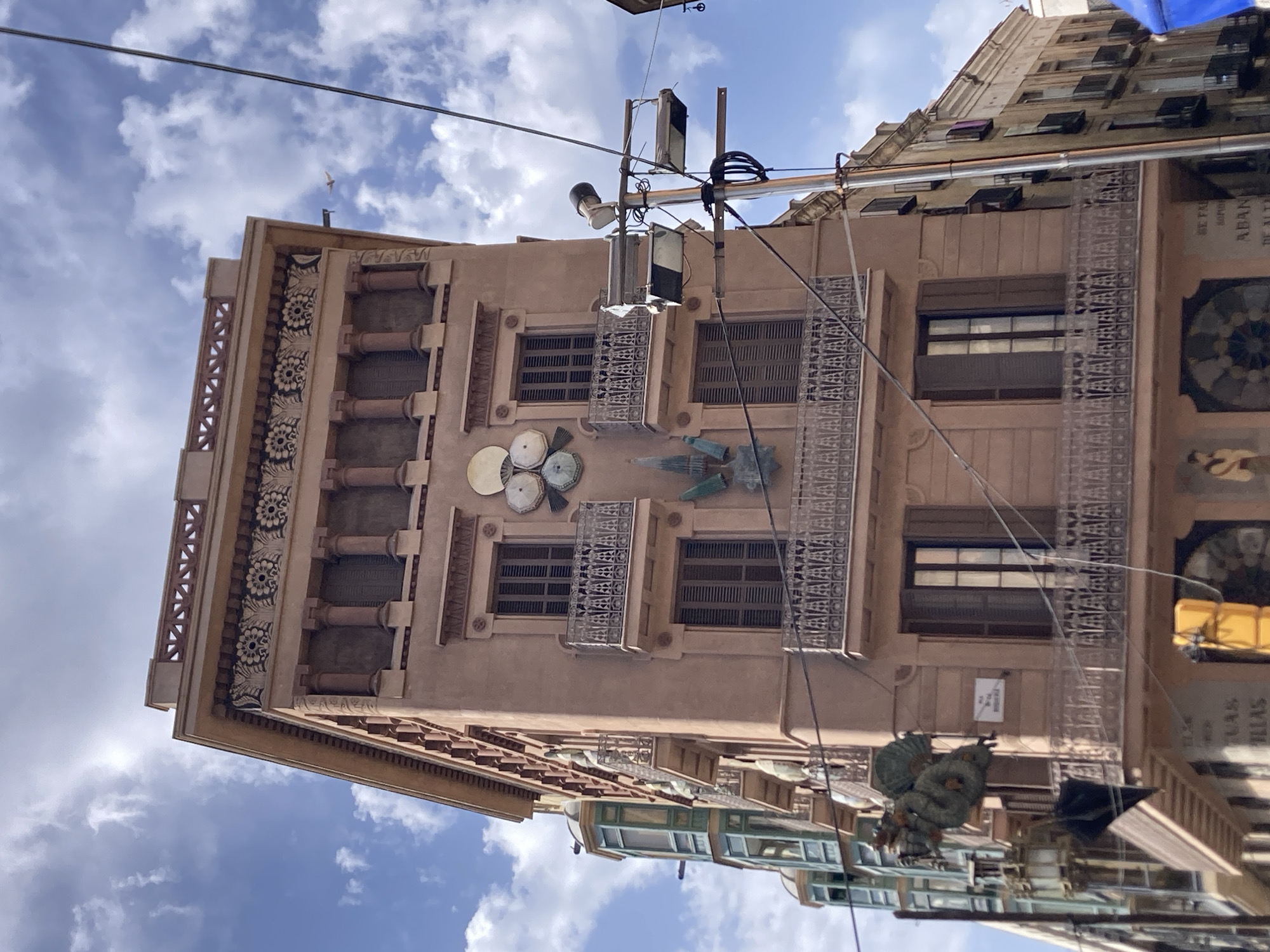 A building with a pink facade and ornate architectural details is captured from a low angle, with a blue sky and scattered clouds in the background. The building features multiple stories with windows and balconies. On the facade, there is a circular art installation with four overlapping discs. To the left, a section of a neighboring building with green bay windows is visible. A lamppost with multiple spotlights and wires is positioned to the right of the main building. There are no people clearly visible in the image. The overall scene suggests an urban environment, likely a street in Barcelona, Spain, given the architectural style.