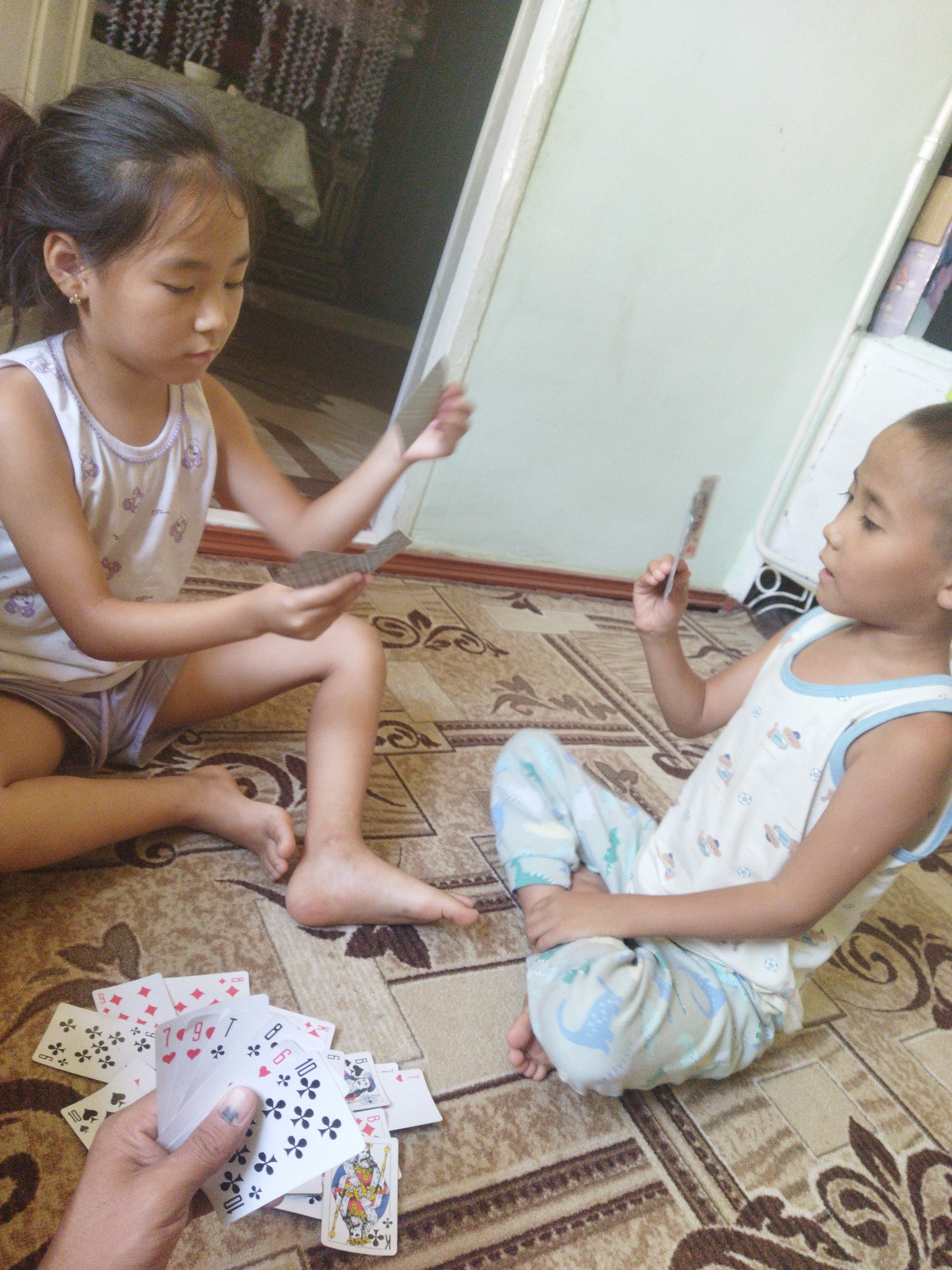 In a room with a patterned carpet, two children are sitting on the floor playing cards. A girl on the left, wearing a white tank top with a pattern of purple bows and grey shorts, holds a fan of cards. A younger child, likely a boy, on the right, wears a white tank top with a pattern of various figures and blue and white dinosaur-patterned pants, and also holds cards. The room appears to be indoors, with a doorway and part of a wall visible in the background. The lighting suggests it is daytime.