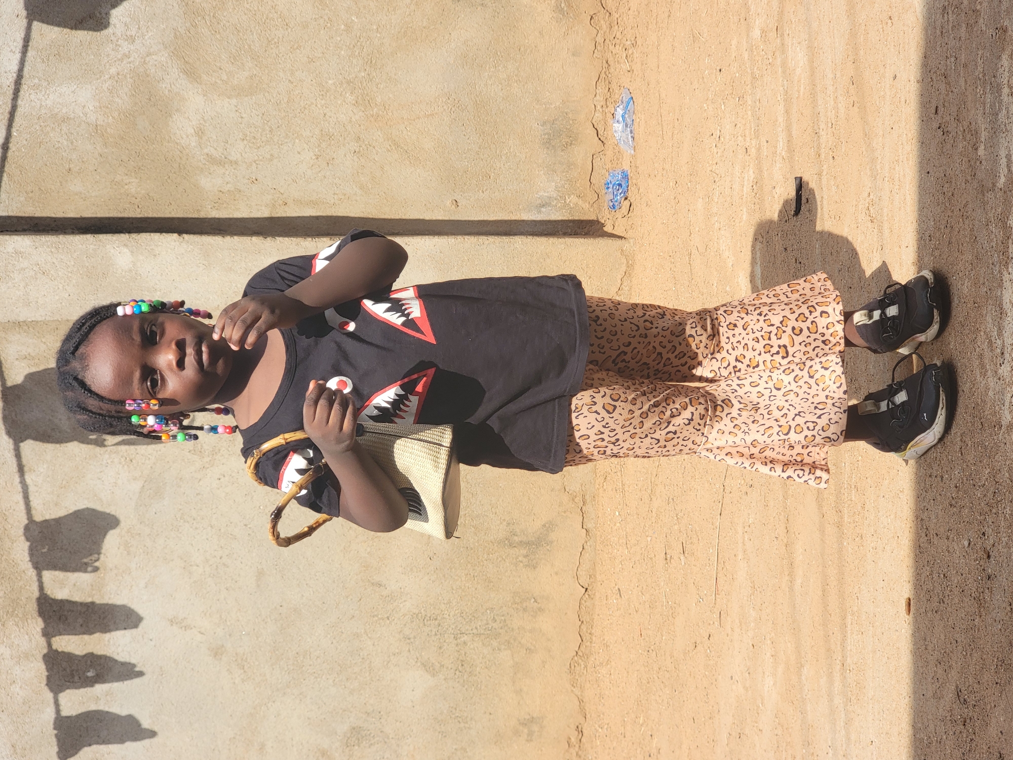 A young girl stands on a dirt and concrete surface adjacent to a concrete wall. She wears a black t-shirt with shark-like teeth designs, leopard-print pants, and black sneakers. Her hair is styled with colorful beads. She is holding a straw handbag. The image was likely taken outside, with strong sunlight casting shadows. The location is Sabon Tasha, Nigeria.