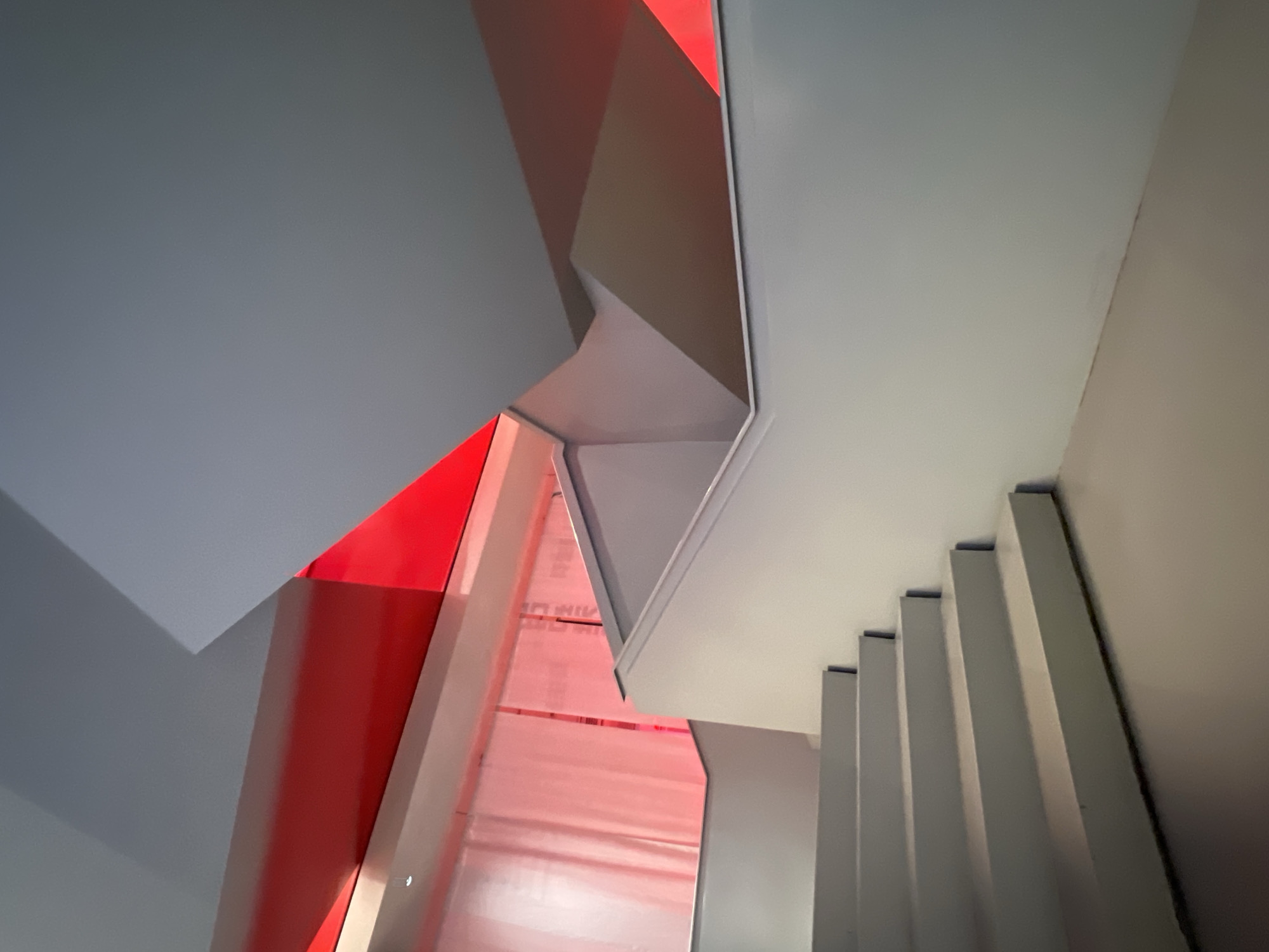 A low-angle shot of a stairwell in Saint Petersburg, Russia, shows steps ascending along the right side of the frame. The steps appear to be made of white or light-colored material. Overhead, sections of the ceiling or architectural elements create a dynamic composition of lines and angles. The lighting is varied, with portions of the scene illuminated in shades of red, and other areas in cooler white or gray tones. The design is modern, featuring sharp edges and geometric shapes.