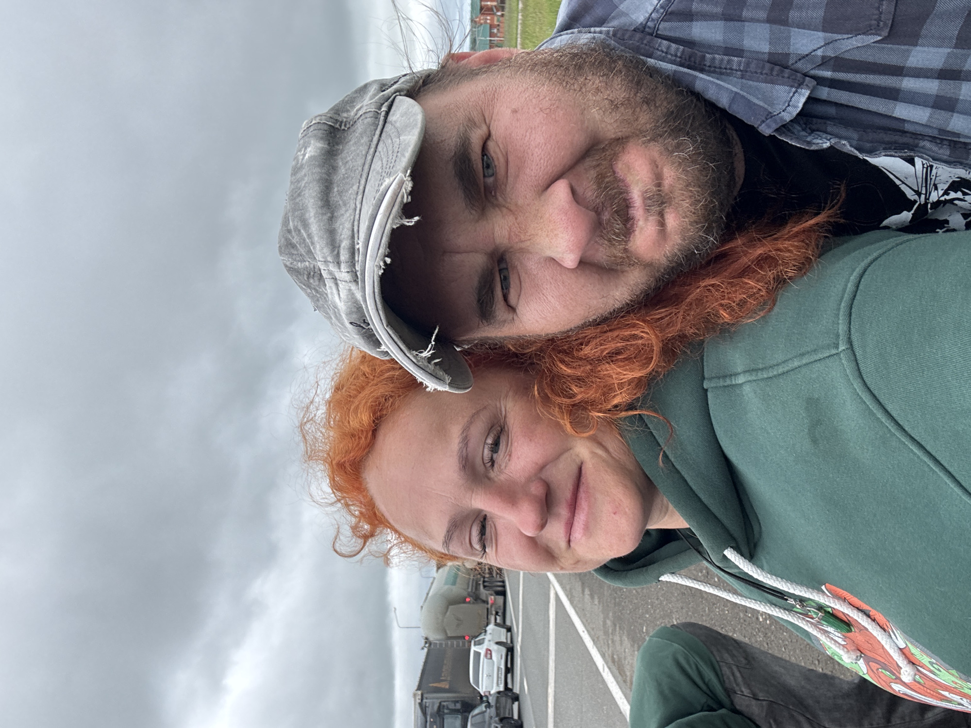 This is a selfie taken by two individuals outdoors, with a road and vehicles visible in the background. The woman in the foreground on the left has vibrant red, curly hair and is wearing a green hooded sweatshirt with white drawstrings. She is smiling. To her right, a man is also looking at the camera, wearing a grey, distressed baseball cap and a blue and grey plaid shirt. He has a beard and is making a grimacing expression. The environment appears to be a roadside in Alnashi, Russia, with traffic including trucks and cars on a highway. The sky is overcast and grey.