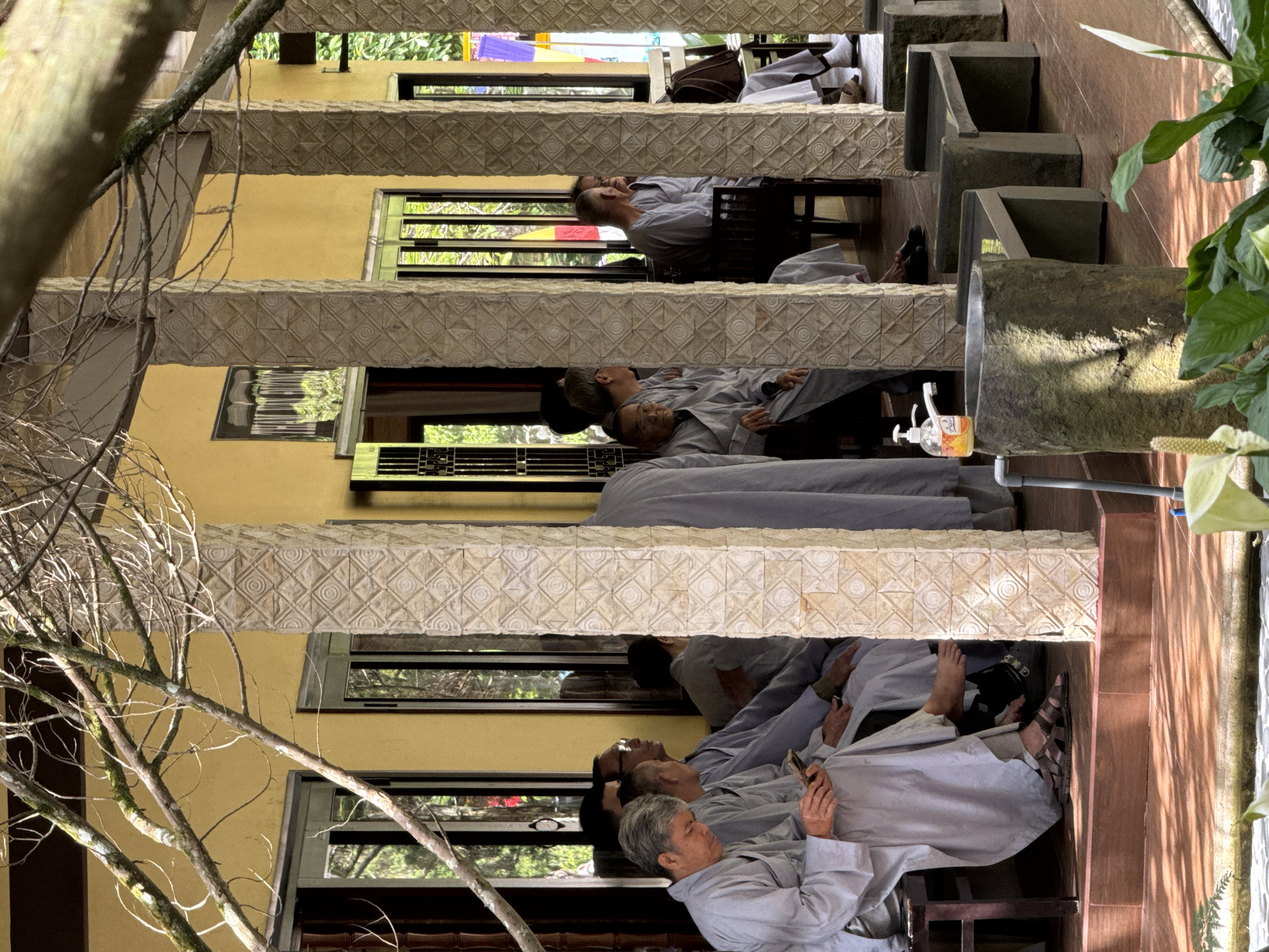 The image captures a detailed scene on a covered veranda in Da Lat, Vietnam. Multiple individuals, predominantly males of varying ages, are seated on dark wooden benches. All are dressed in simple, light grey monastic or devotional robes. In the foreground, an older man in a grey robe is using a smartphone, looking down at the device. Other robed figures are seated along the yellow wall, some conversing, others observing, or resting. The veranda features light yellow walls and substantial rectangular pillars carved with a repeating geometric pattern of squares and circles. Bare, moss-covered tree branches extend across the top and upper left of the frame, partially obscuring the building. Sunlight from the right casts distinct shadows on the brown tiled floor. In the lower right foreground, a stone basin serves as a wash station, equipped with a faucet and a pump-style hand sanitizer dispenser. Adjacent to it are several dark grey, minimalist stone benches. Green foliage and white peace lily flowers are visible in the immediate foreground. Through windows and openings in the background, lush greenery and faintly visible colorful prayer flags (red, yellow, blue) can be discerned.