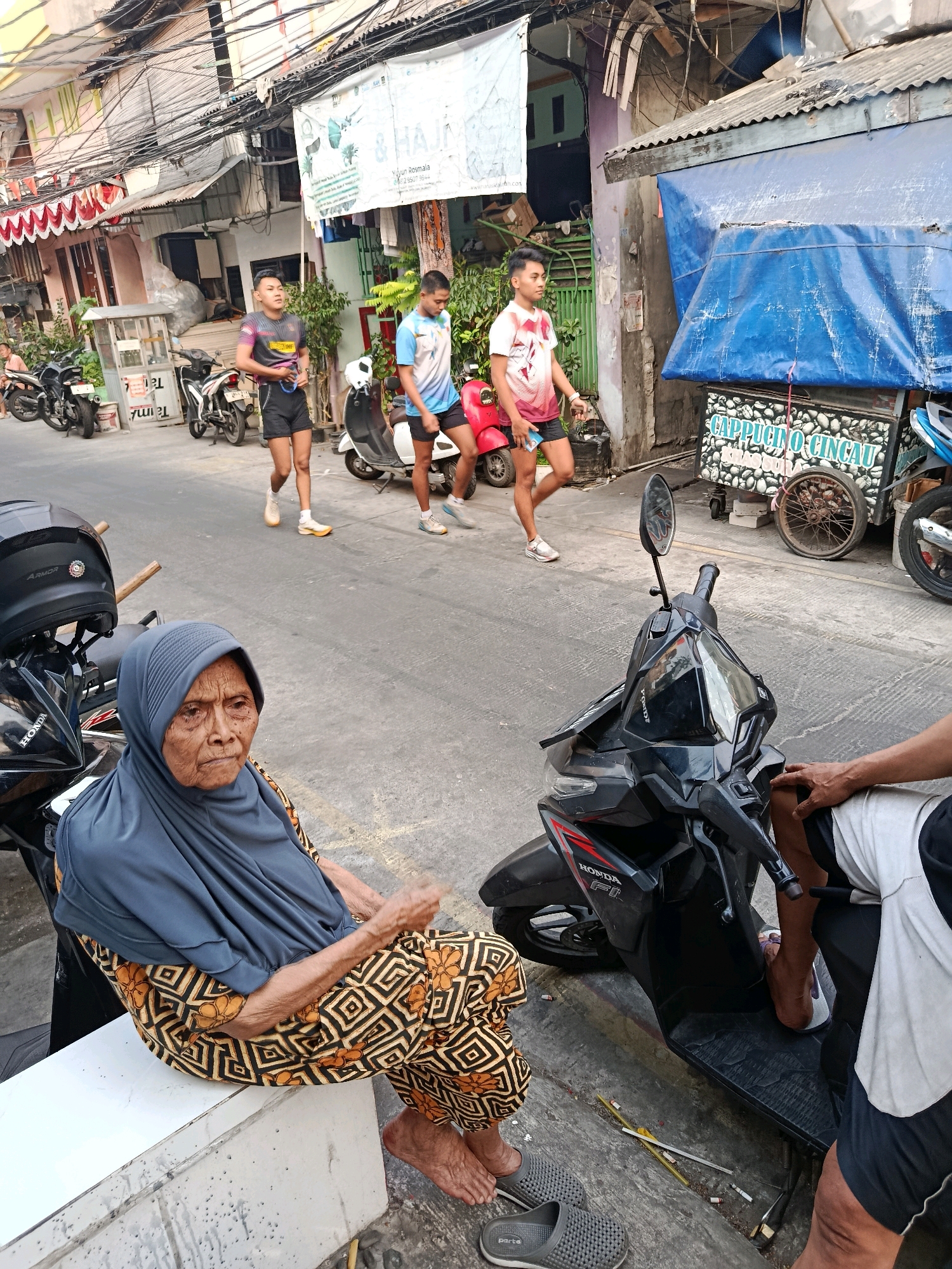 An elderly Indonesian woman wearing a grey hijab and a patterned sarong sits on a white concrete ledge on the right side of a street in North Jakarta, Indonesia. She is looking to her right with a somber expression. Beside her, to the left, is a black motorcycle with a black helmet on its handlebars. In the background, three young Indonesian men in athletic wear are walking down the street. One of them is holding a blue and white water bottle. They are passing a food cart with a sign that reads "CAPPPUCCINO CINCAU" and several parked motorcycles. The street is lined with buildings, some with corrugated metal roofs and colorful awnings. Overhead, a dense network of electrical wires is visible. The ground is paved with asphalt and concrete.