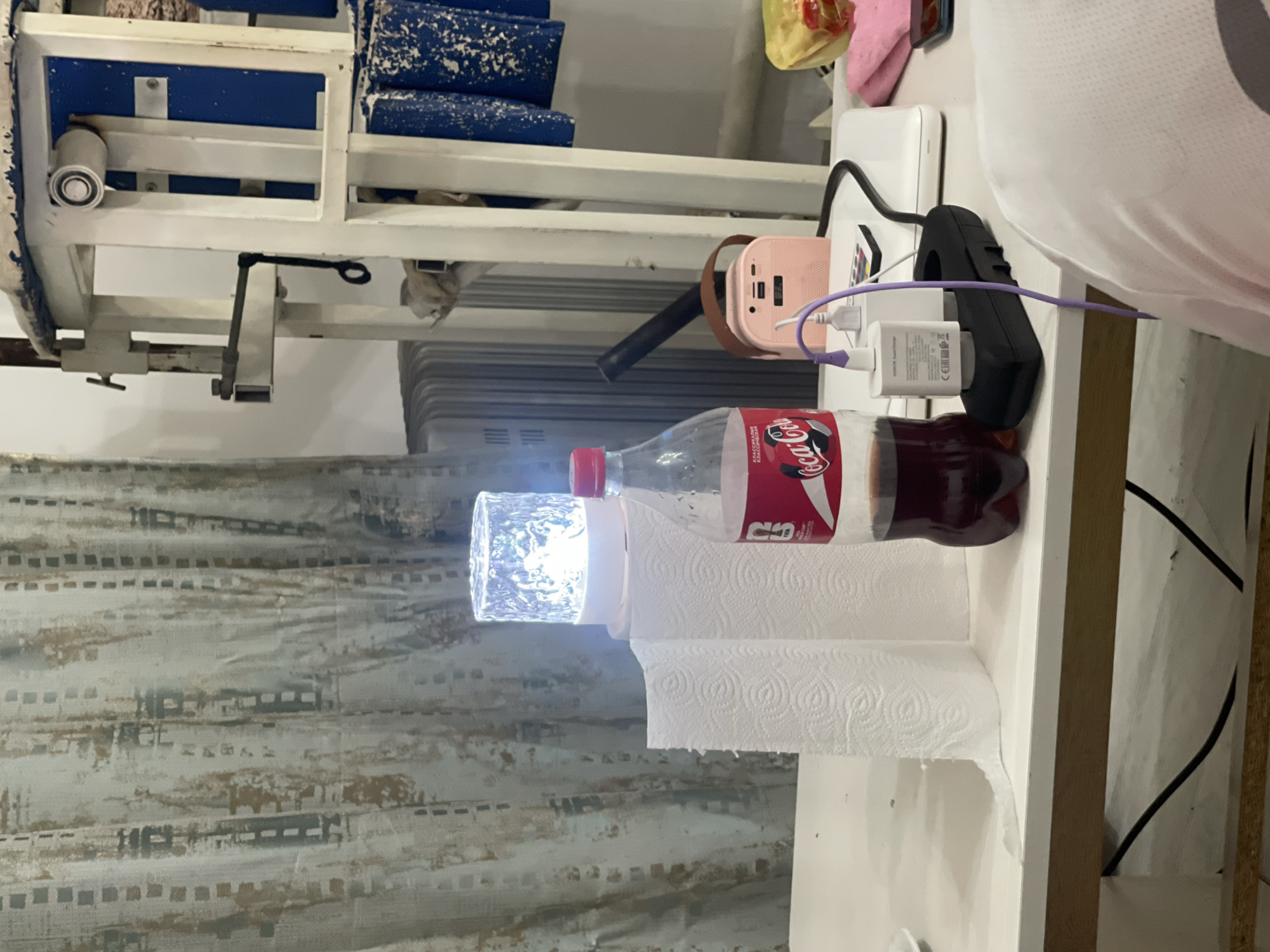The image shows a cluttered table with various items. In the foreground, a person's shoulder is visible, wearing a white, textured shirt. To the left, two rolls of paper towels are standing upright. Behind them, a clear glass object with a light inside illuminates, giving off a bright white glow. Next to the paper towels is a large plastic bottle of Coca-Cola, filled with a dark liquid.
Further back on the table, several electronic devices are present. A pink portable speaker with a brown handle is visible, connected to a power source. Next to it is a white charging brick and a black power strip with a purple cable plugged into it. A white laptop or tablet is also on the table. To the far right, a pink cloth and a smartphone are visible.
The background features a patterned curtain with vertical stripes and abstract shapes in neutral tones. Behind the table, part of a white and blue metal structure with various components is visible, along with a radiator. The overall scene appears to be indoors, in a domestic or workspace setting. There is no discernible information about the location being in Bishkek City, Kyrgyzstan.