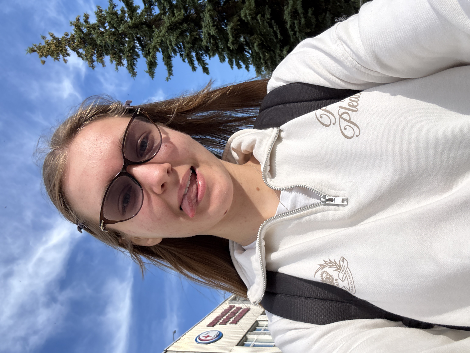 A young woman, wearing glasses and a white zip-up hoodie, sticks her tongue out at the camera while posing for a selfie. She has brown hair, tied back with a clip. In the background, a green evergreen tree stands against a bright blue sky with wispy white clouds. To the left of the tree, a building with a sign that has a red cross and the numbers "629" is visible. The woman is carrying a black backpack. The scene appears to be outdoors in a city setting. Based on the provided metadata, the location is Chaikovsky, Russia.