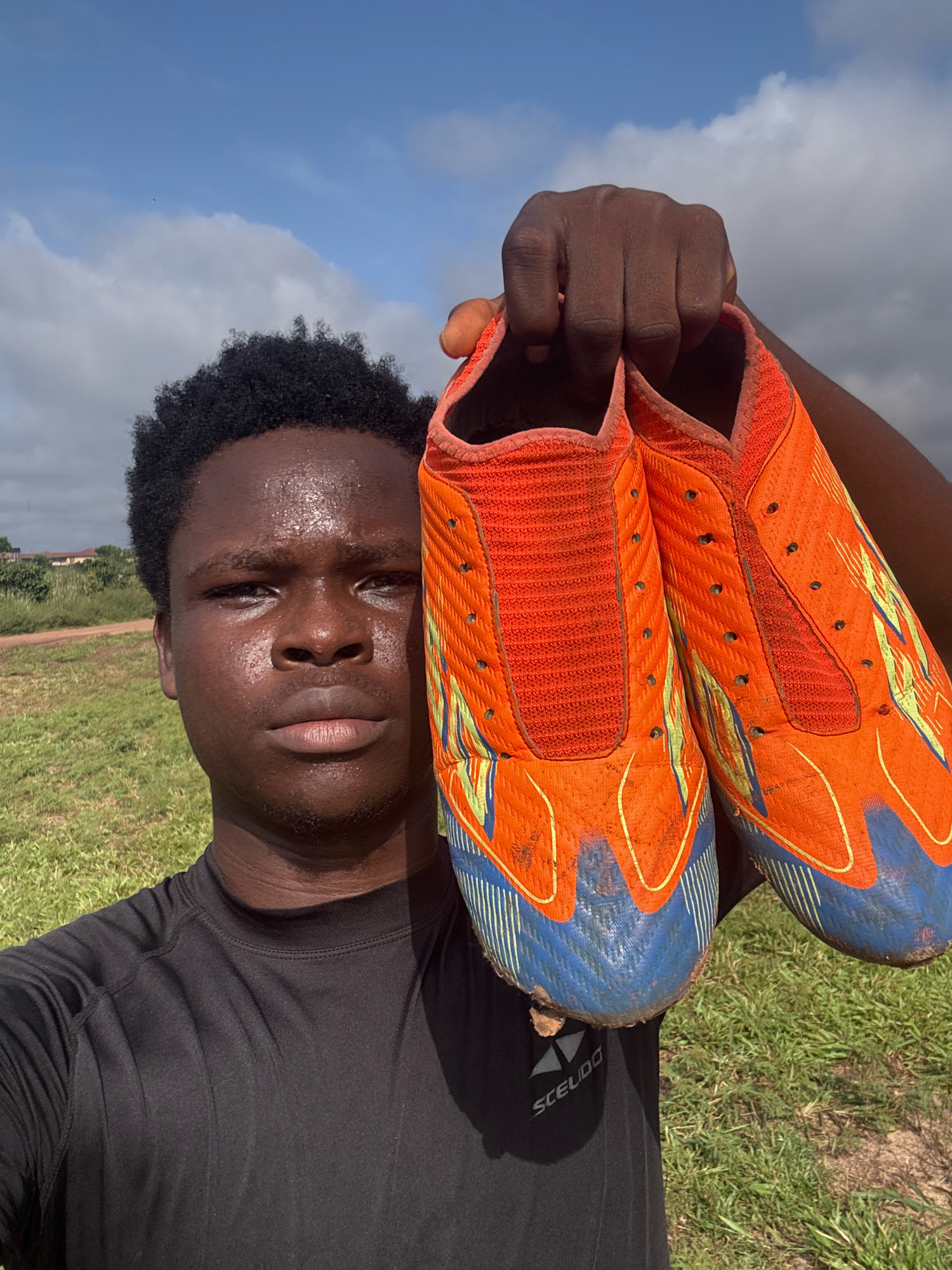 A young Black male, appearing to be in his late teens or early twenties, stands outdoors, holding up a pair of bright orange football cleats. He is wearing a black t-shirt. The cleats are dirty, suggesting recent use on a field. The background is a grassy field under a partly cloudy sky, with some residential buildings visible in the distance. The overall scene appears to be an outdoor recreational area, likely a football pitch, in a rural or semi-urban setting. The lighting suggests it is daytime, possibly late afternoon, given the shadows. The location is identified as Ihiagwa, Nigeria.