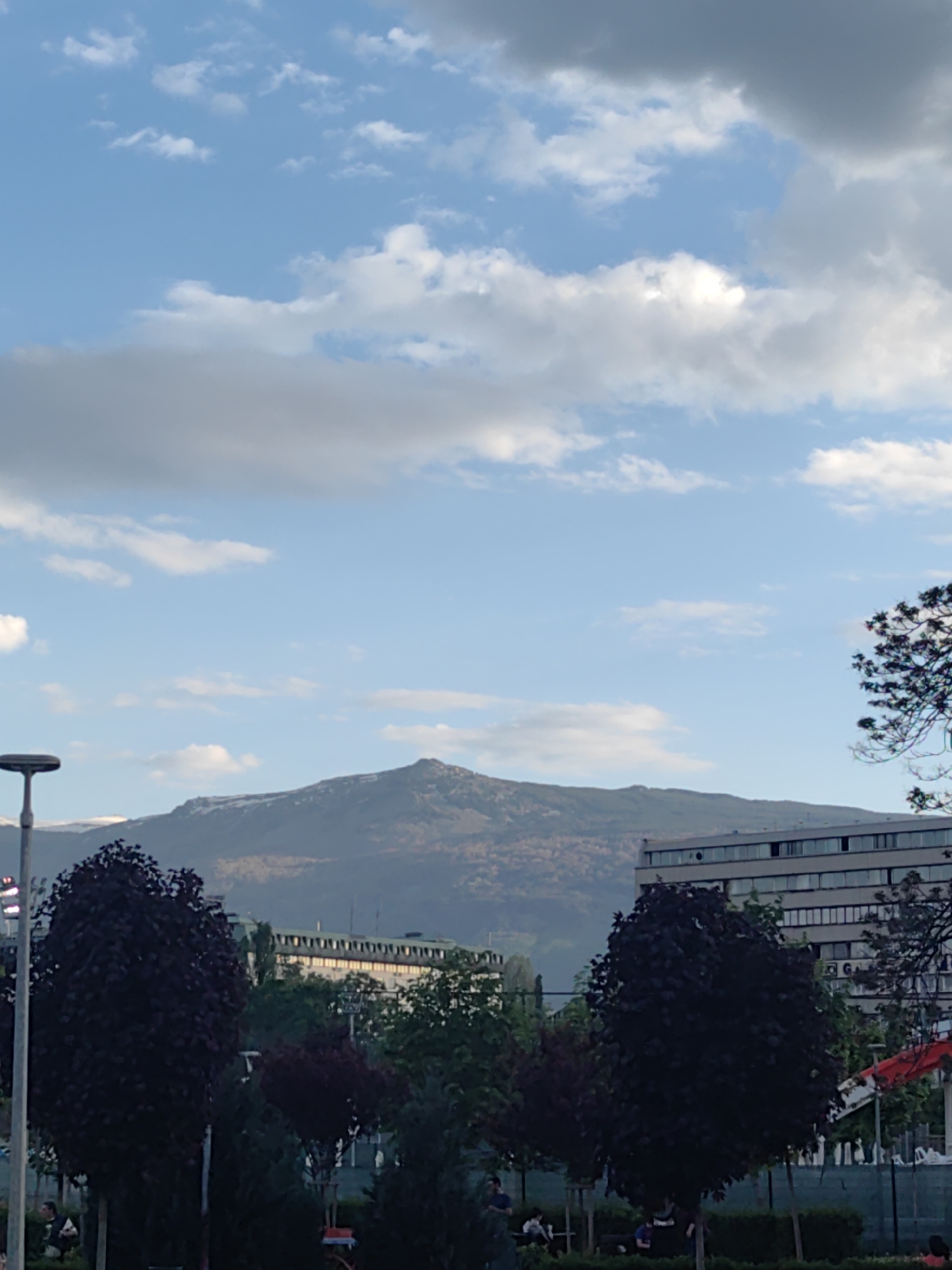 The image shows a view towards Vitosha Mountain in Sofia, Bulgaria. In the foreground is a park with trees (some with dark purple foliage), lampposts, and a few people sitting on benches. In the midground, there are various buildings, including a long, multi-story building with many windows. The background is dominated by Vitosha Mountain, with some snow visible near the peak. The sky is blue with scattered white clouds and some darker, grey clouds.