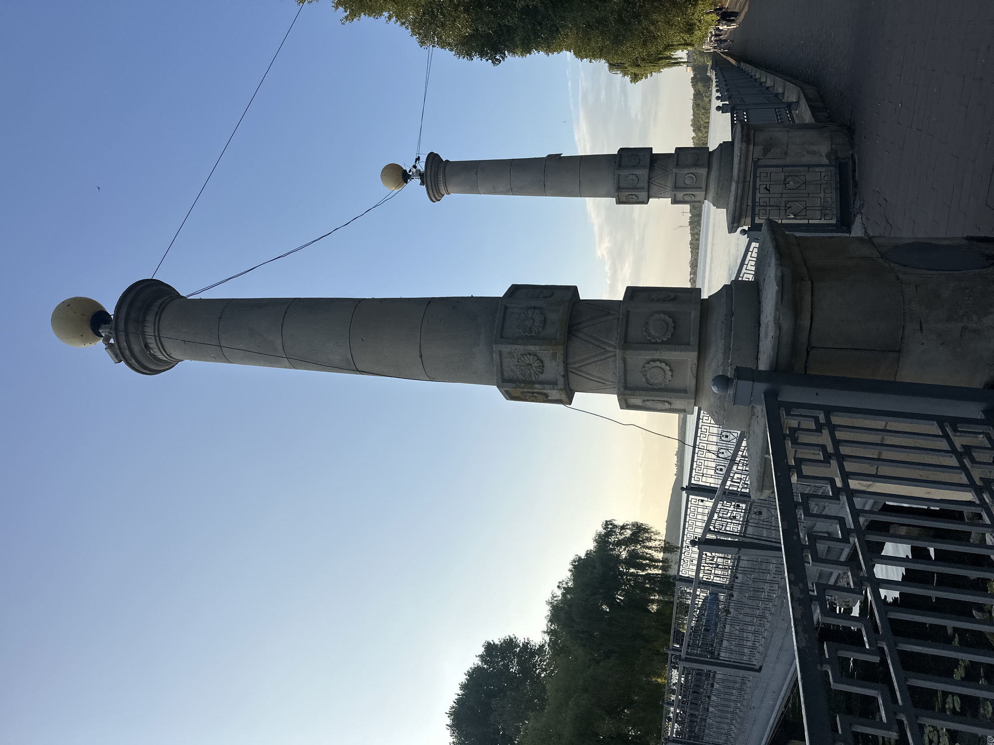 The image captures a scene along the Ternopil Lake embankment in Ternopil, Ukraine. In the foreground, two ornate stone pillars with spherical lamps on top flank a paved walkway. The pillars feature decorative geometric patterns and floral motifs. A gray metal railing separates the walkway from the lake, and a bridge with a similar railing is visible to the left, arching over the water.
Beyond the railing, the calm surface of Ternopil Lake reflects the clear blue sky, which transitions to a lighter hue near the horizon, suggesting late afternoon or early evening. Lush green trees line the embankment on the left and right sides of the image, with weeping willows creating a softer texture on the left.
A few individuals can be seen walking along the embankment in the distance on the right, their figures small against the vastness of the landscape. The overall atmosphere is serene and picturesque, typical of a park or recreational area by the water. The presence of the pillars and the bridge suggest a well-maintained public space designed for leisure and appreciation of the natural scenery.