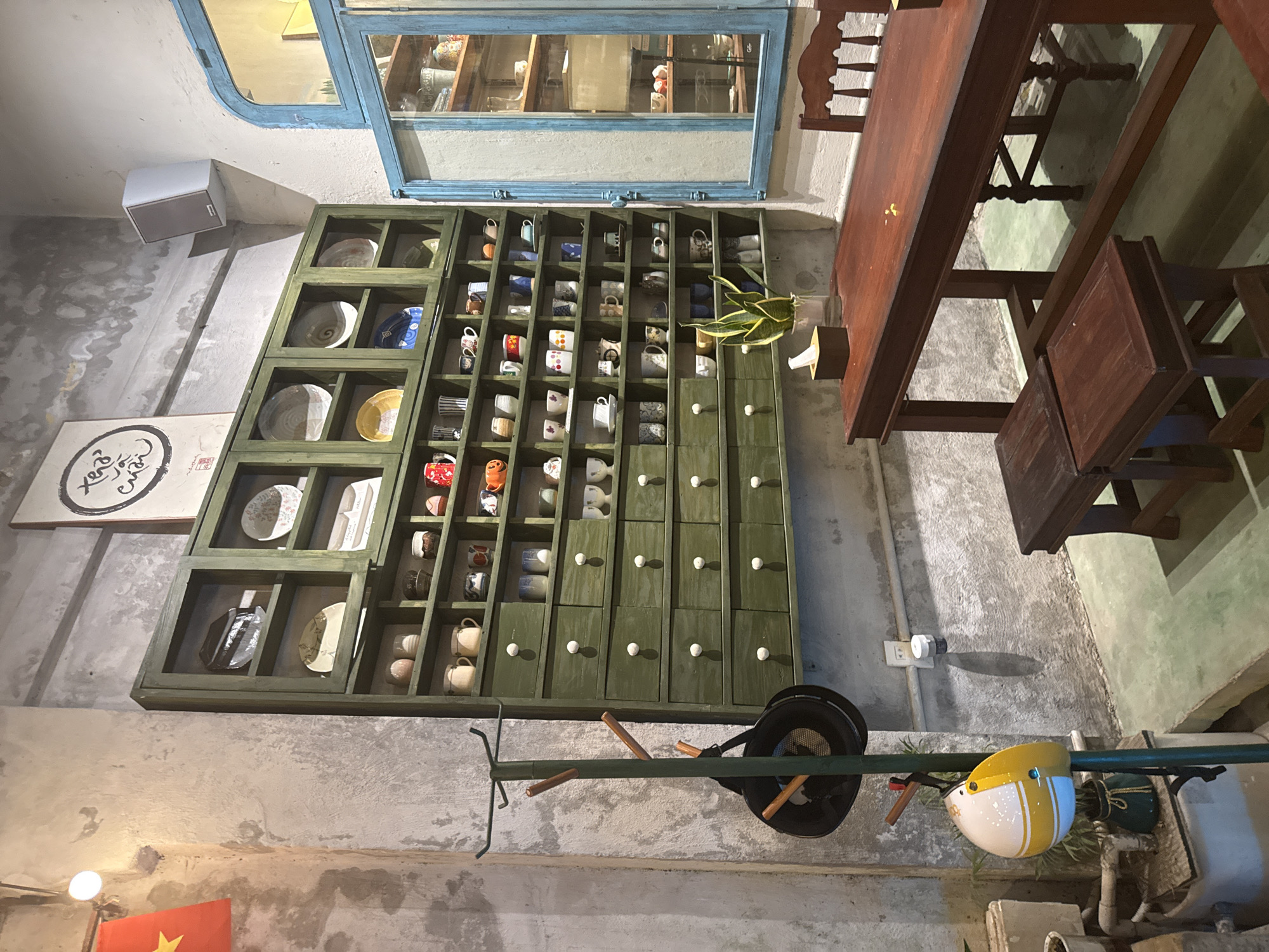 This image appears to be taken in a cafe or restaurant in Đà Nẵng, Vietnam.
The interior has a rustic, possibly vintage, vibe, with exposed concrete walls. A large green cabinet or display case dominates one wall, filled with shelves and drawers. The shelves hold an assortment of ceramic items, mostly cups and plates, likely for sale or display. Below the cabinet hangs a metal rod with a motorcycle helmet in white and yellow, and a black wok.
A dark wood table and chairs sit nearby, typical of cafe seating. Above the cabinet, there is a window displaying additional ceramic items. The scene is lit with natural light and possibly a warm artificial light source. A small flag from Vietnam is located in the bottom left of the image.