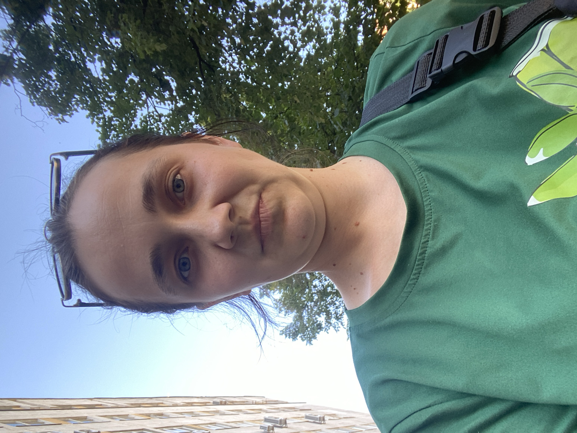 A medium shot captures a woman from the chest up, posing for a selfie outdoors. She has light skin, blue eyes, and dark hair pulled back, with glasses perched on top of her head. Her expression is neutral. She is wearing a green t-shirt with a design of green leaves on the front, and a black strap from a bag is visible across her right shoulder. In the background, a tall, light-colored building with visible windows stands on the left, and leafy green trees are on the right, set against a bright blue sky. The lighting suggests it is daytime.