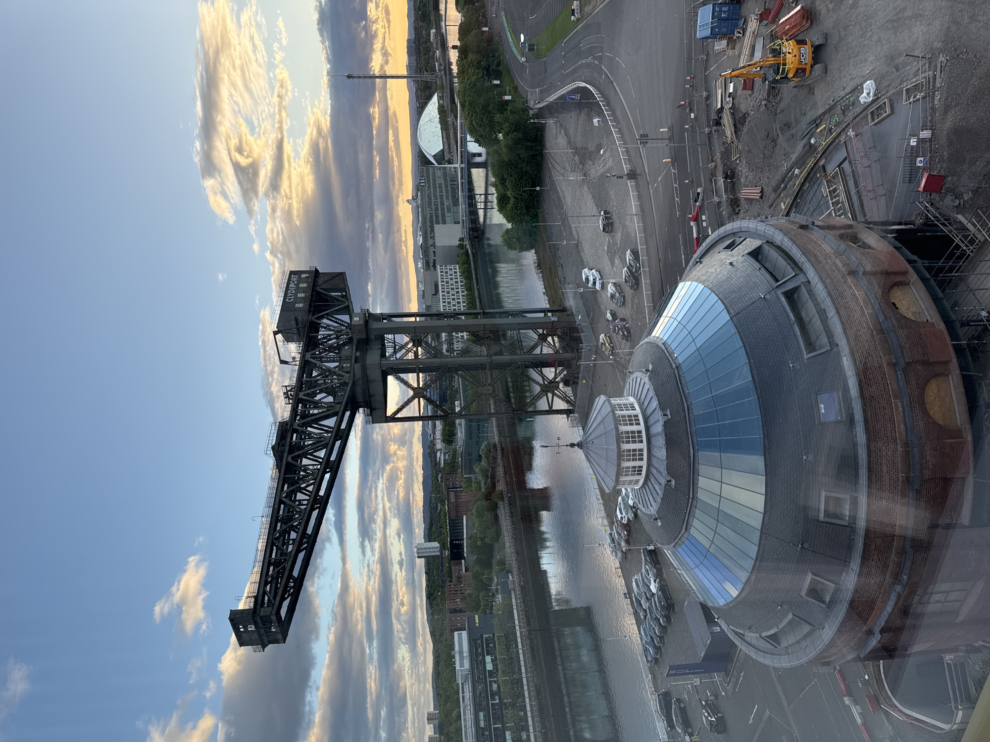 The image captures a view in Glasgow, United Kingdom, overlooking the River Clyde. The Finnieston Crane, a prominent landmark, dominates the left side of the frame. To the right of the crane is the Glasgow Science Centre, notable for its curved, metal roof. Below these structures lies the River Clyde, reflecting the colors of the evening sky. Cars are parked near the Science Centre. A road and other buildings are visible in the background. The sky displays a mix of blue and cloudy conditions, with a hint of sunset hues. Construction equipment is visible near the Science Centre.