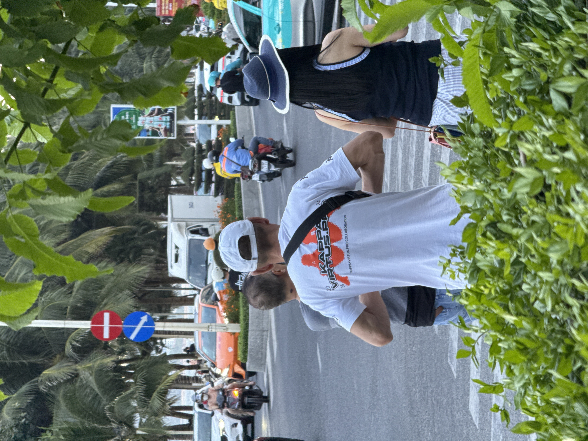 A group of people are standing and walking on a street in Nha Trang Ward, Vietnam, with palm trees and lush green foliage in the foreground and background. In the mid-ground, a man in a white t-shirt with a red and black logo is visible from behind, along with another person in a grey shirt. A woman in a blue hat and a black top with white stripes is standing next to them. Cars, motorcycles, and traffic signs are present on the street, indicating a busy urban environment. The overall scene depicts a typical street view in a tropical city.