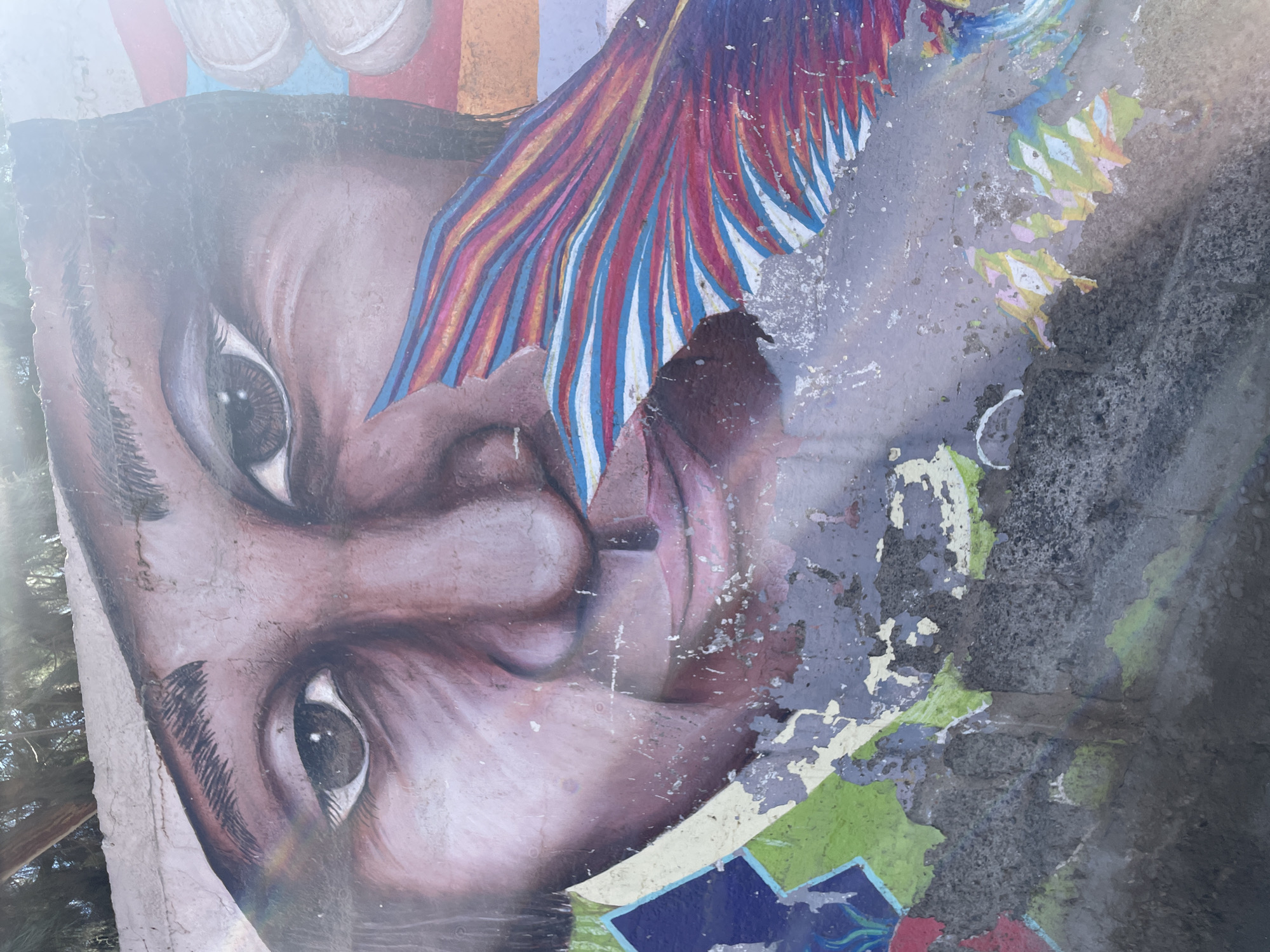 A mural of a person's face is depicted on a damaged wall. The face, rendered in earthy tones, is partially obscured by peeling paint and the textured surface of the wall. The eyes are open and looking forward, with one eye prominently featured. Streaks of colorful paint, resembling feathers or flowing fabric, emanate from the right side of the face. Below the face, the wall is further damaged, with large sections of paint missing, revealing the rough concrete beneath. Geometric shapes in blue and green are visible on the lower left side of the mural. The upper part of the image is bright, suggesting sunlight is illuminating the scene. The mural is located on a wall in an outdoor environment, with trees visible in the background.